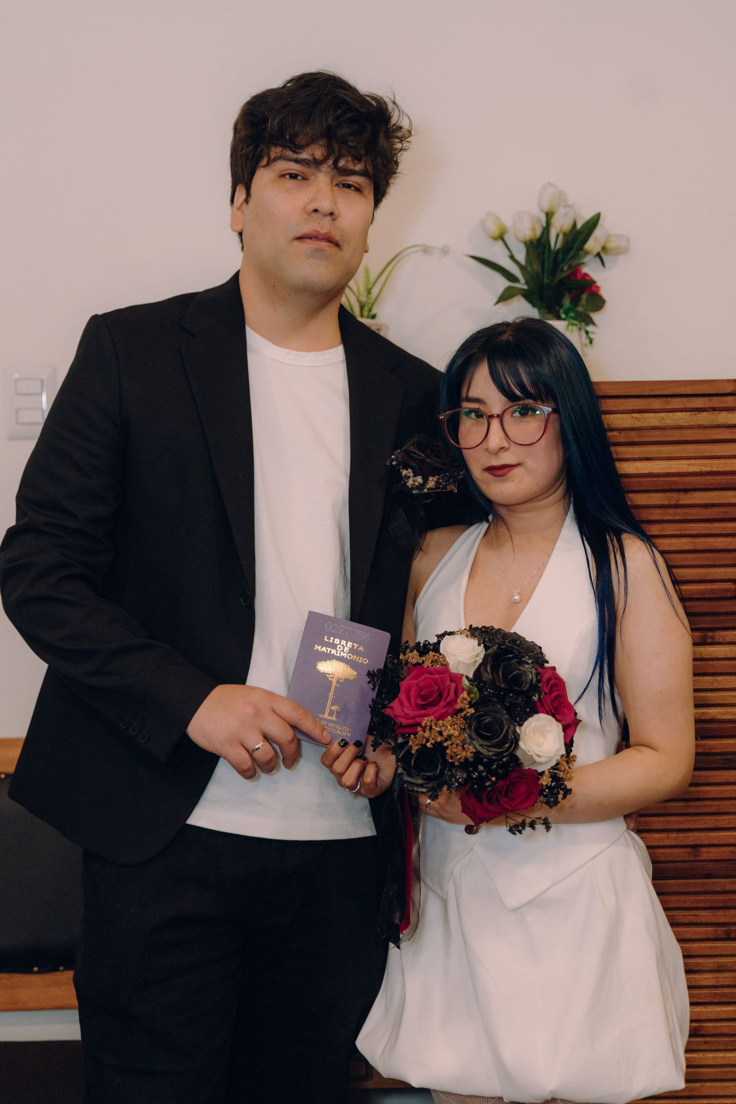 Bride with blue hair holds dark floral bouquet beside groom in black suit at civil wedding.