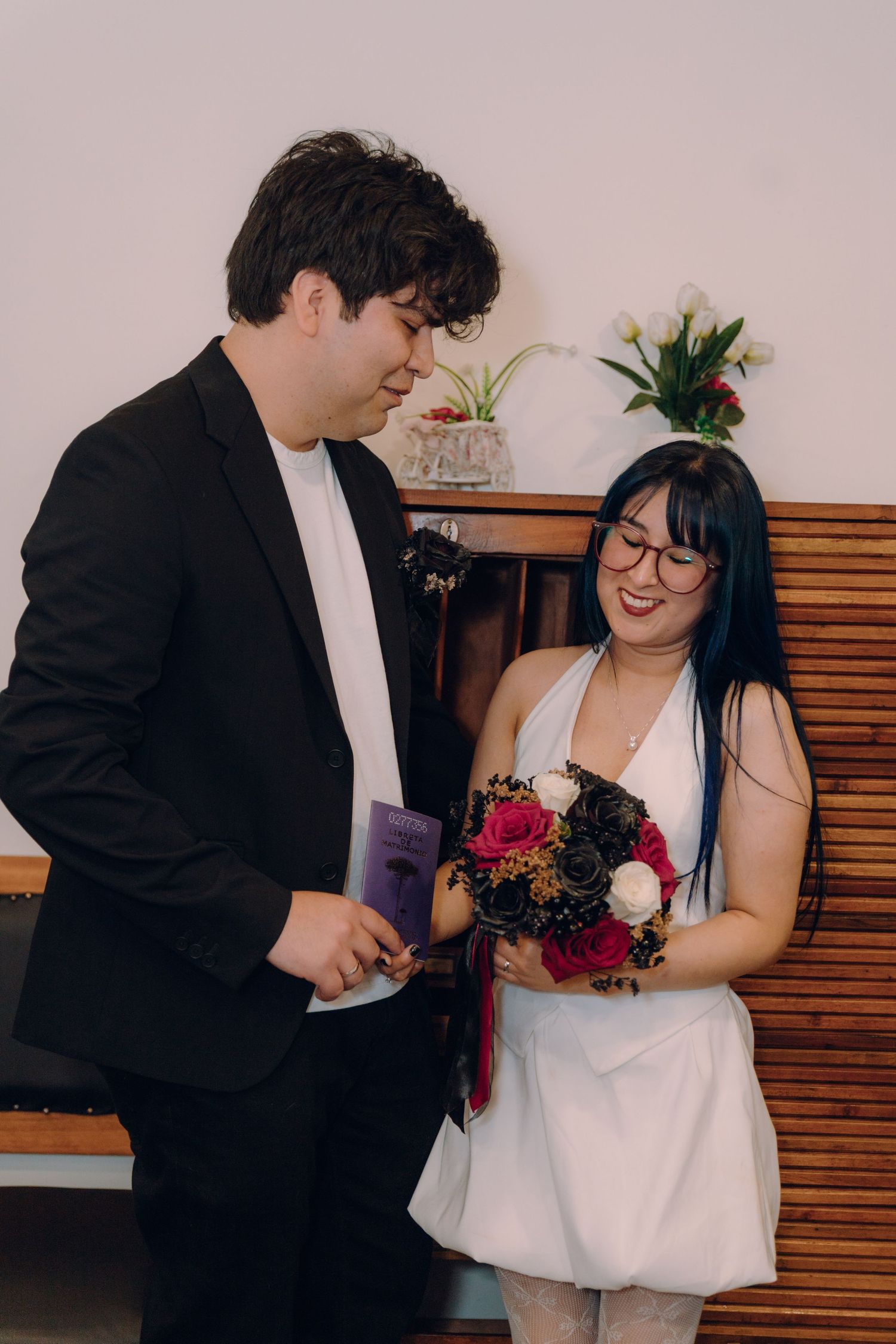 Smiling bride in white dress holds floral bouquet while groom in black suit looks on lovingly.