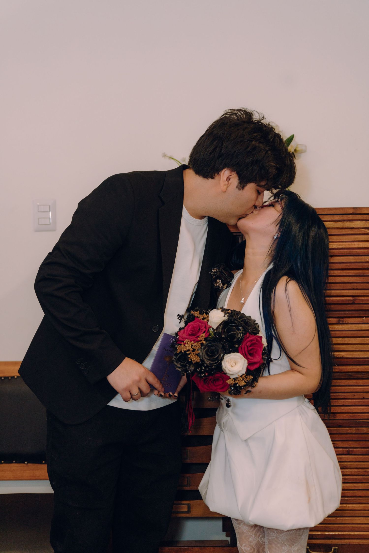 Newlyweds share a romantic kiss, bride holding dark floral bouquet in white dress.