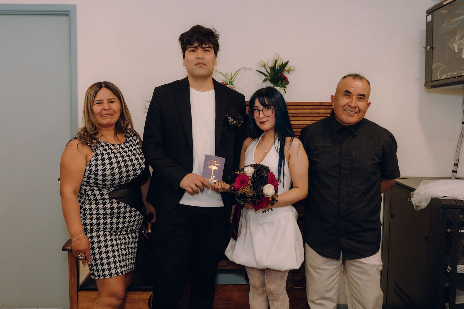 Newlyweds pose with two guests, bride holding red roses bouquet in casual wedding celebration.