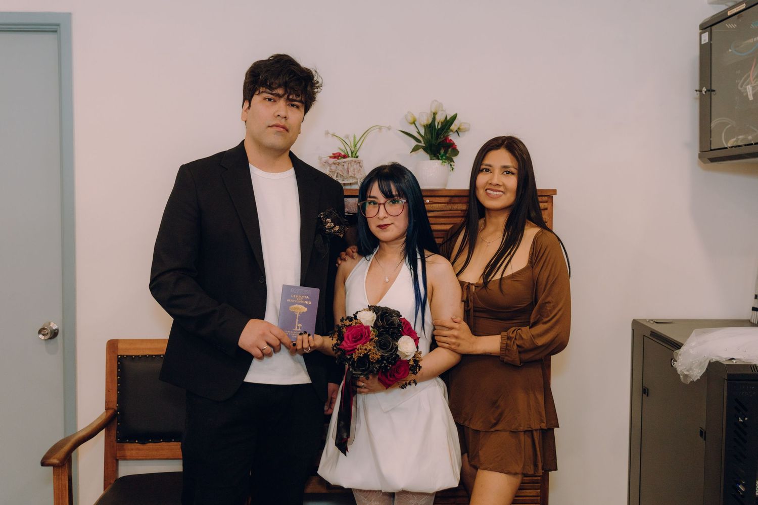 Bride in white dress and groom in black suit pose with female guest at wedding.