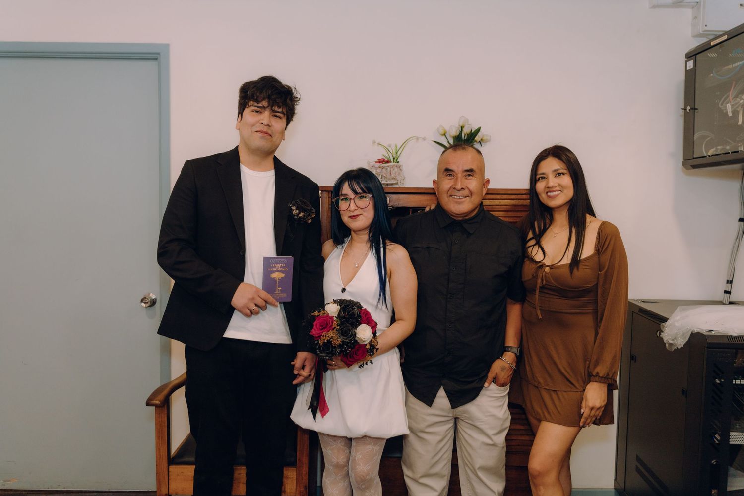 Newlyweds pose with two guests at intimate indoor wedding ceremony, bride holding floral bouquet.