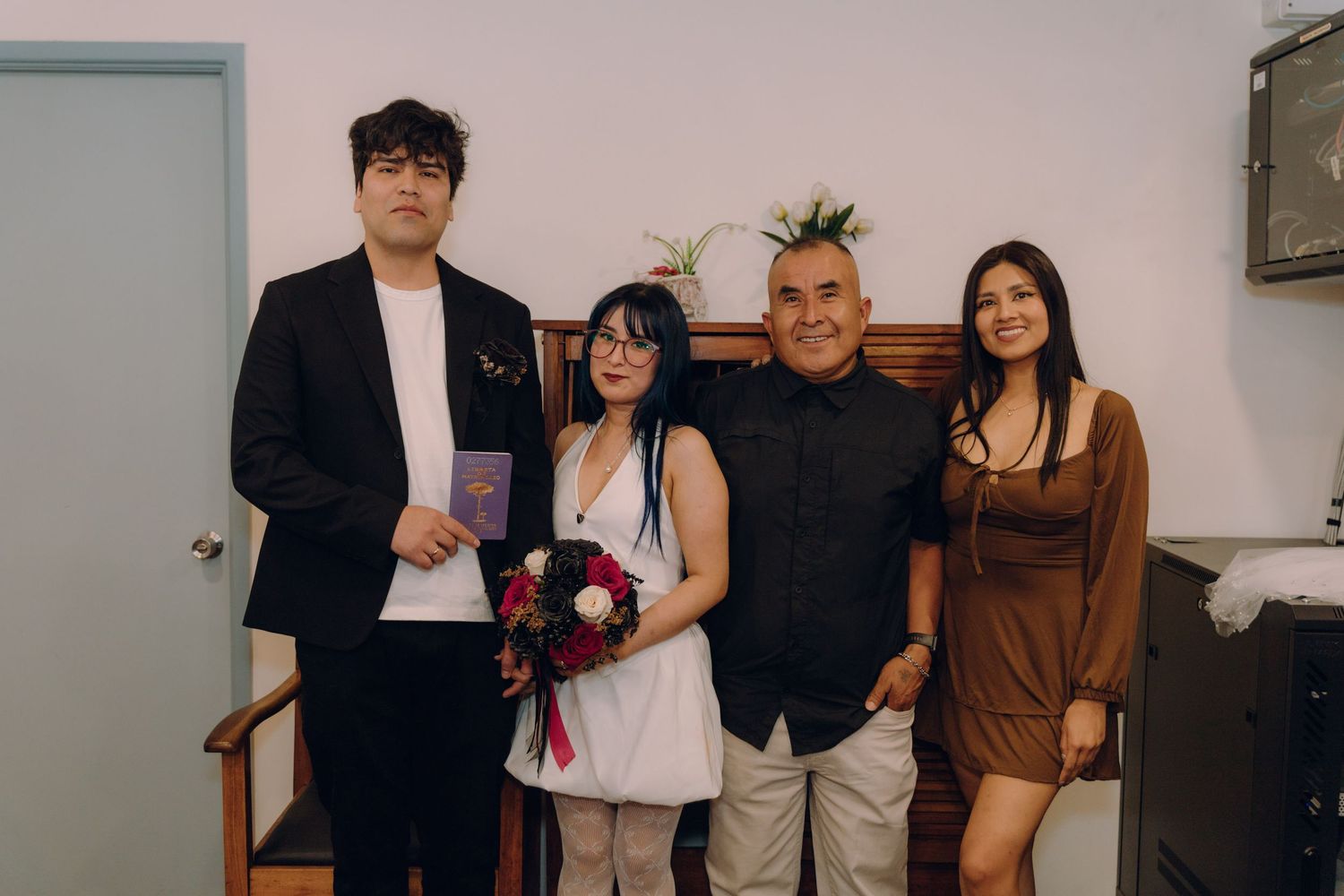 Wedding couple stands with two guests for formal photo at small indoor ceremony.