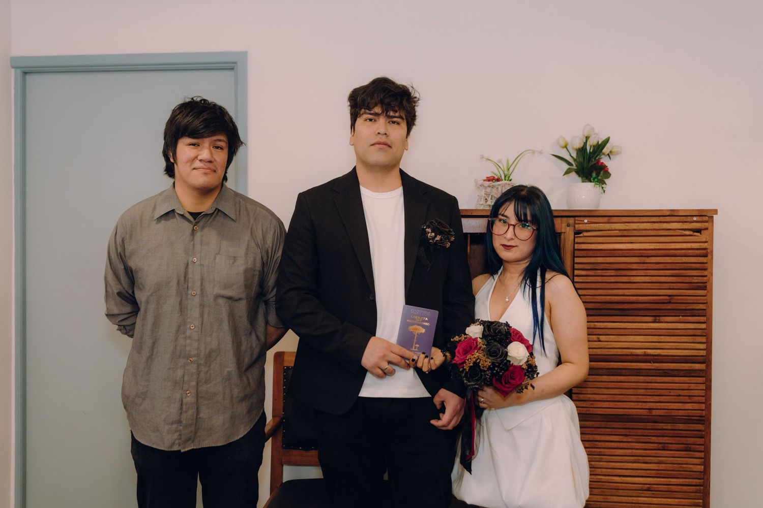 Three people pose formally indoors, man in center holding purple book, woman in white dress holding red bouquet.
