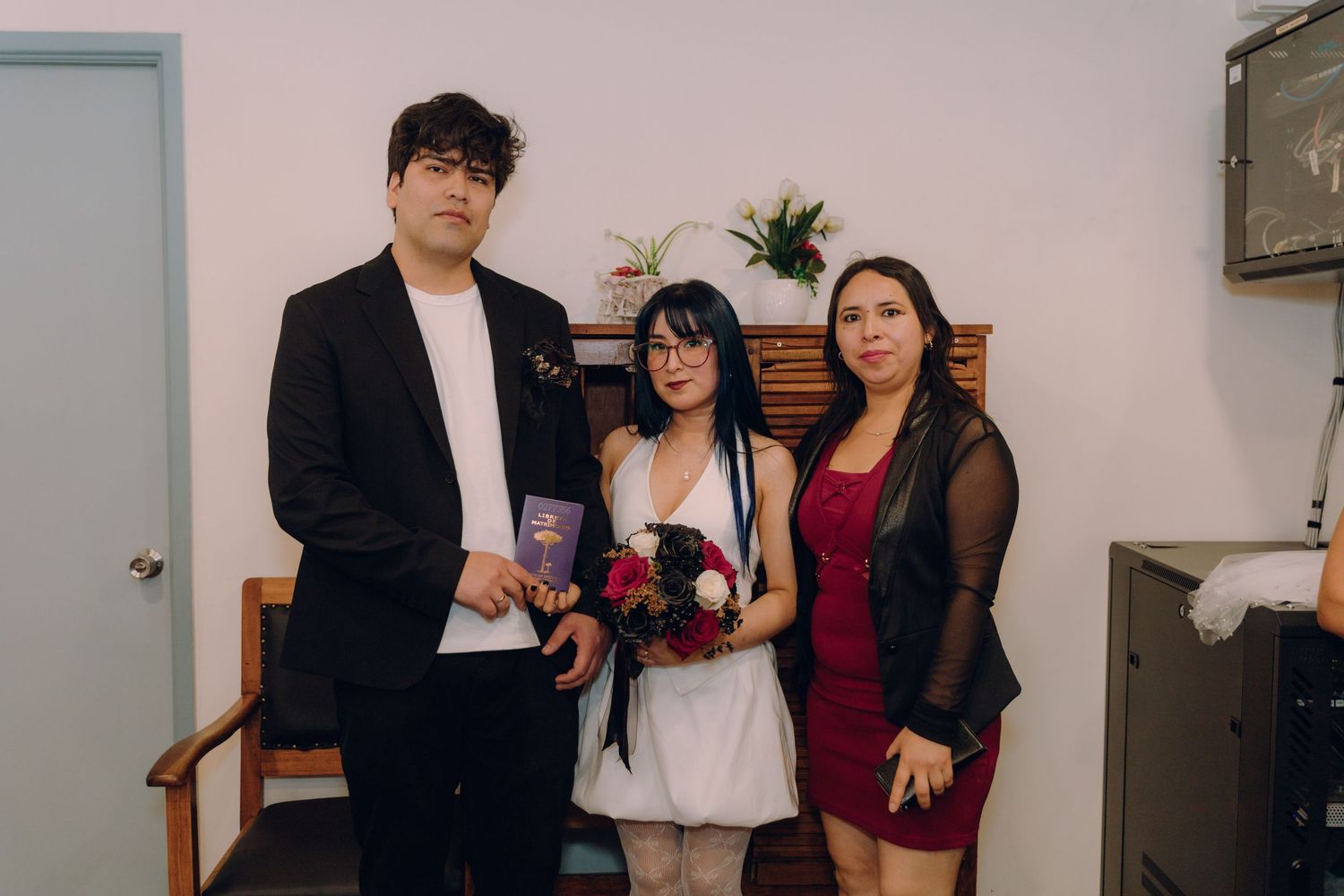 Couple in formal attire poses with older woman, bride holds red and white bouquet indoors.
