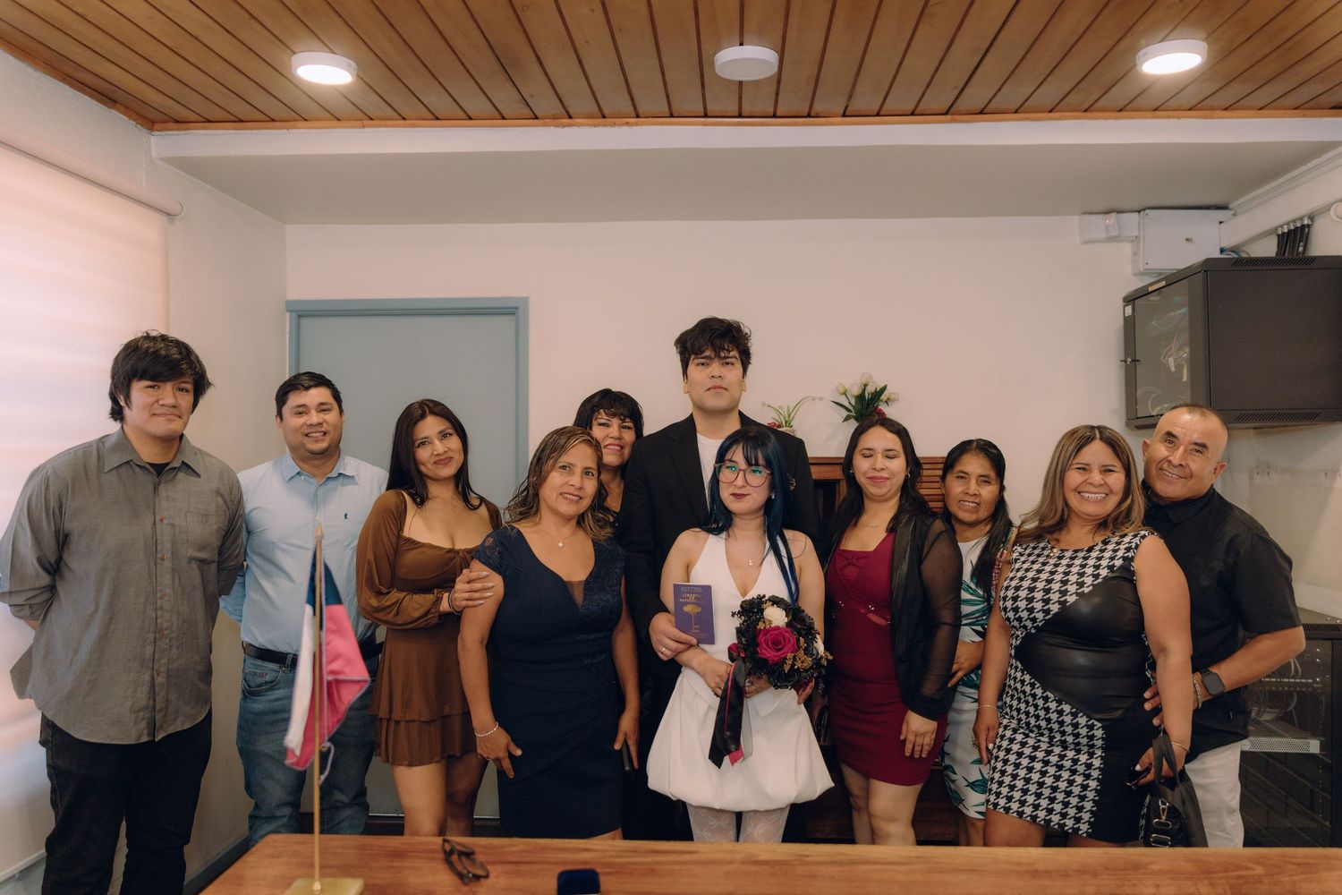 Group of eleven people smile together at an indoor civil wedding ceremony with Chilean flag visible.