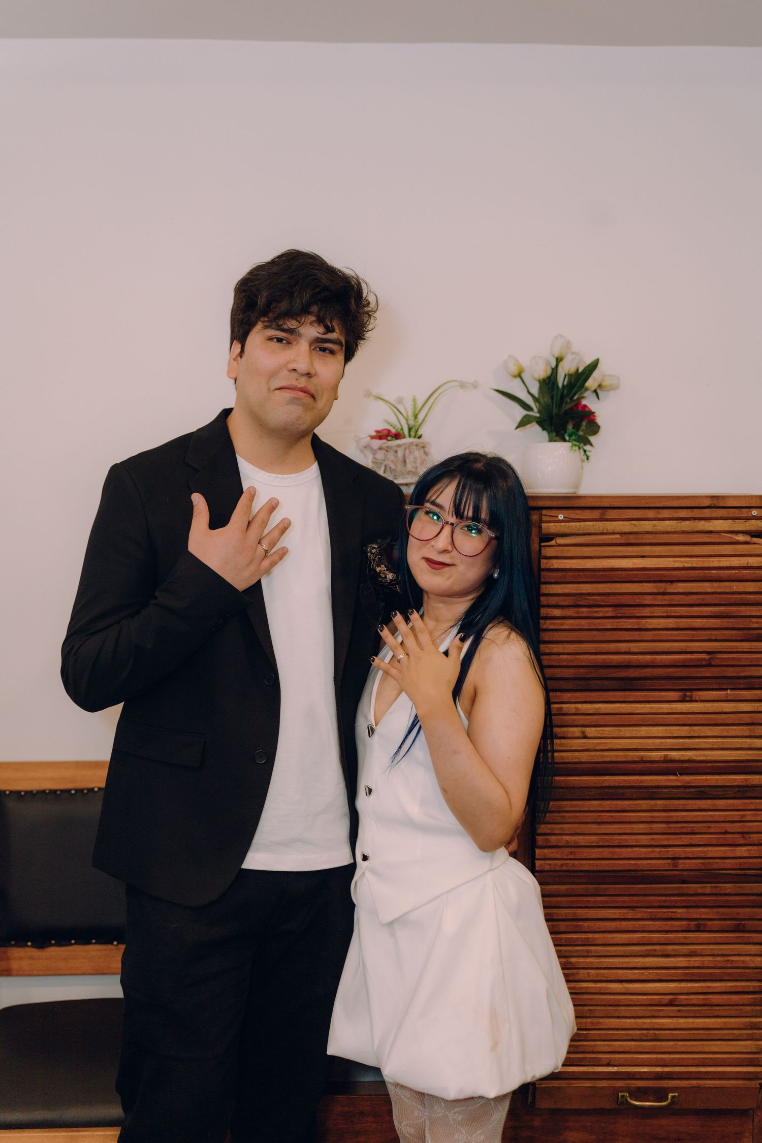 Newlywed couple shows off wedding rings, groom in black suit, bride in white mini dress indoors.