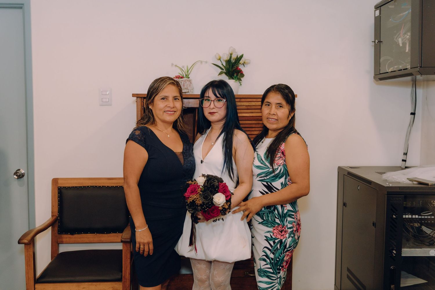 Bride in white dress holds red bouquet while posing with two women at indoor wedding ceremony.