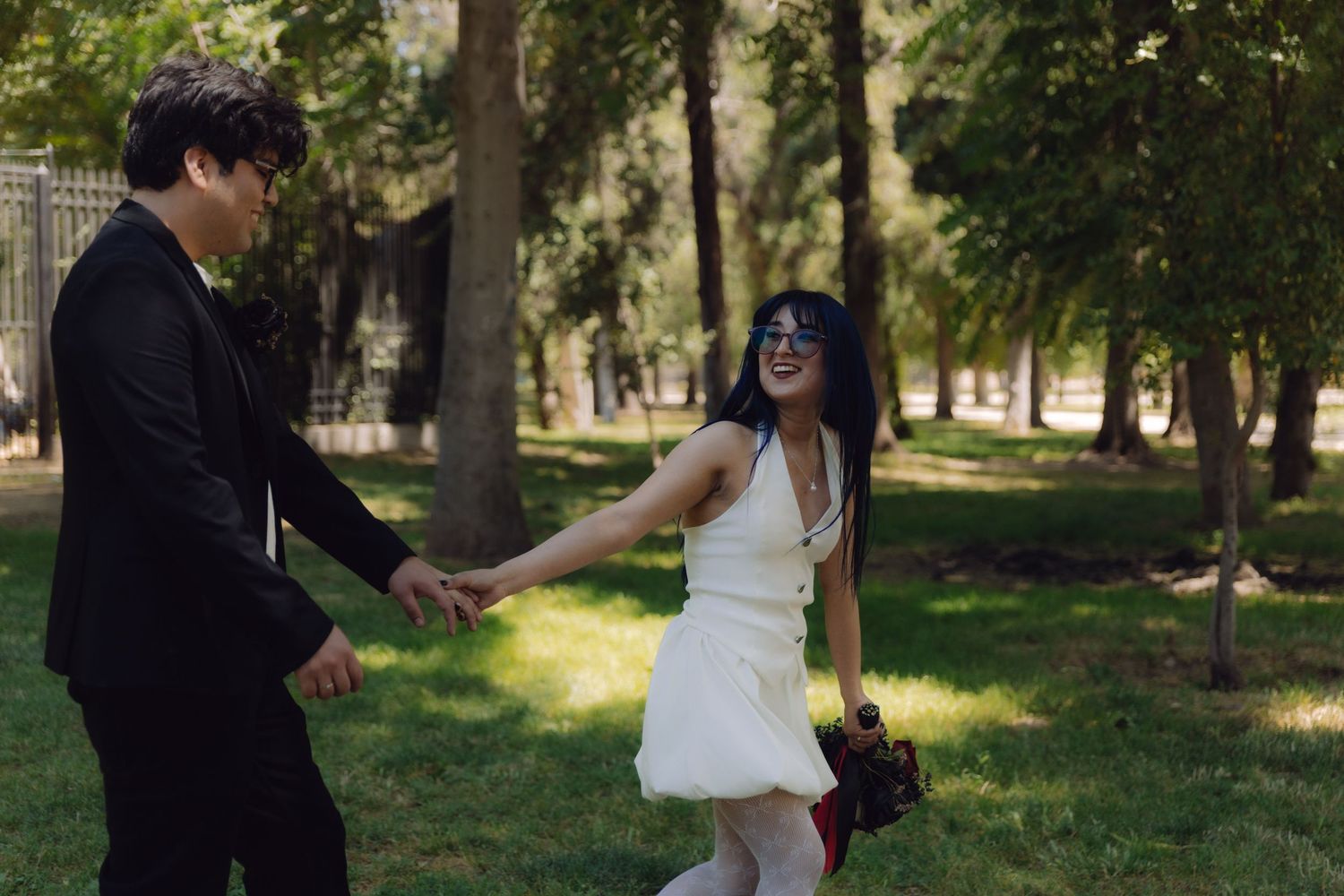Newlywed couple walks hand in hand through sunny green park, bride in white dress and groom in black.