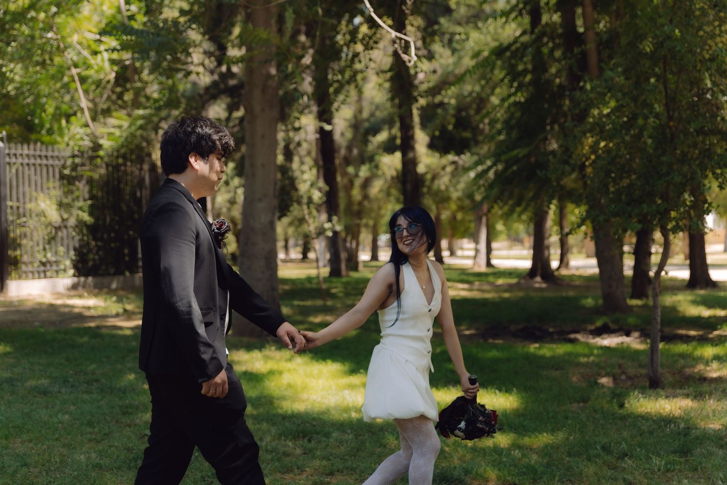 Couple in wedding attire walks holding hands through a sunlit park with tall trees.