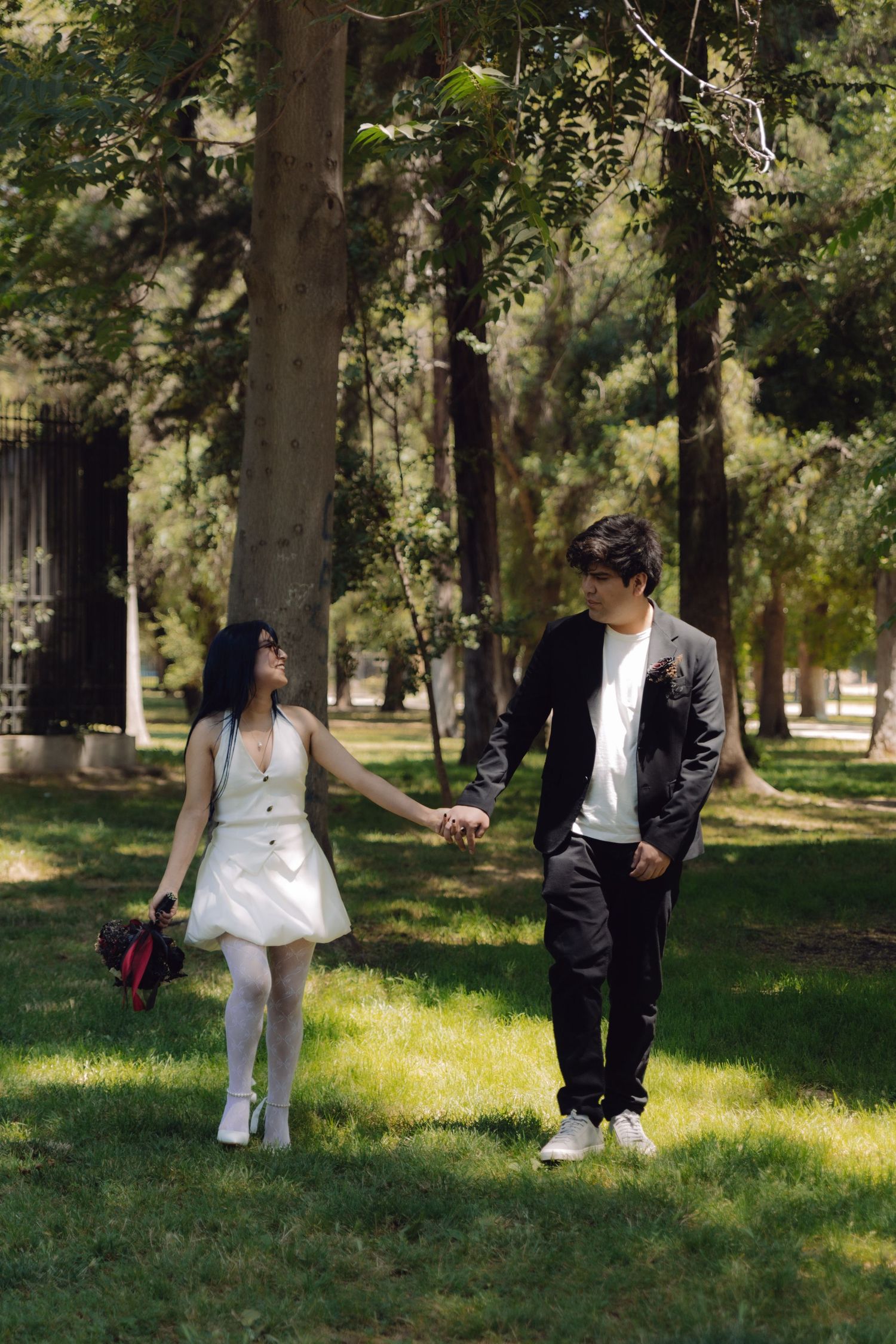 Newlywed couple holds hands and gazes at each other while strolling through a sunny green park.
