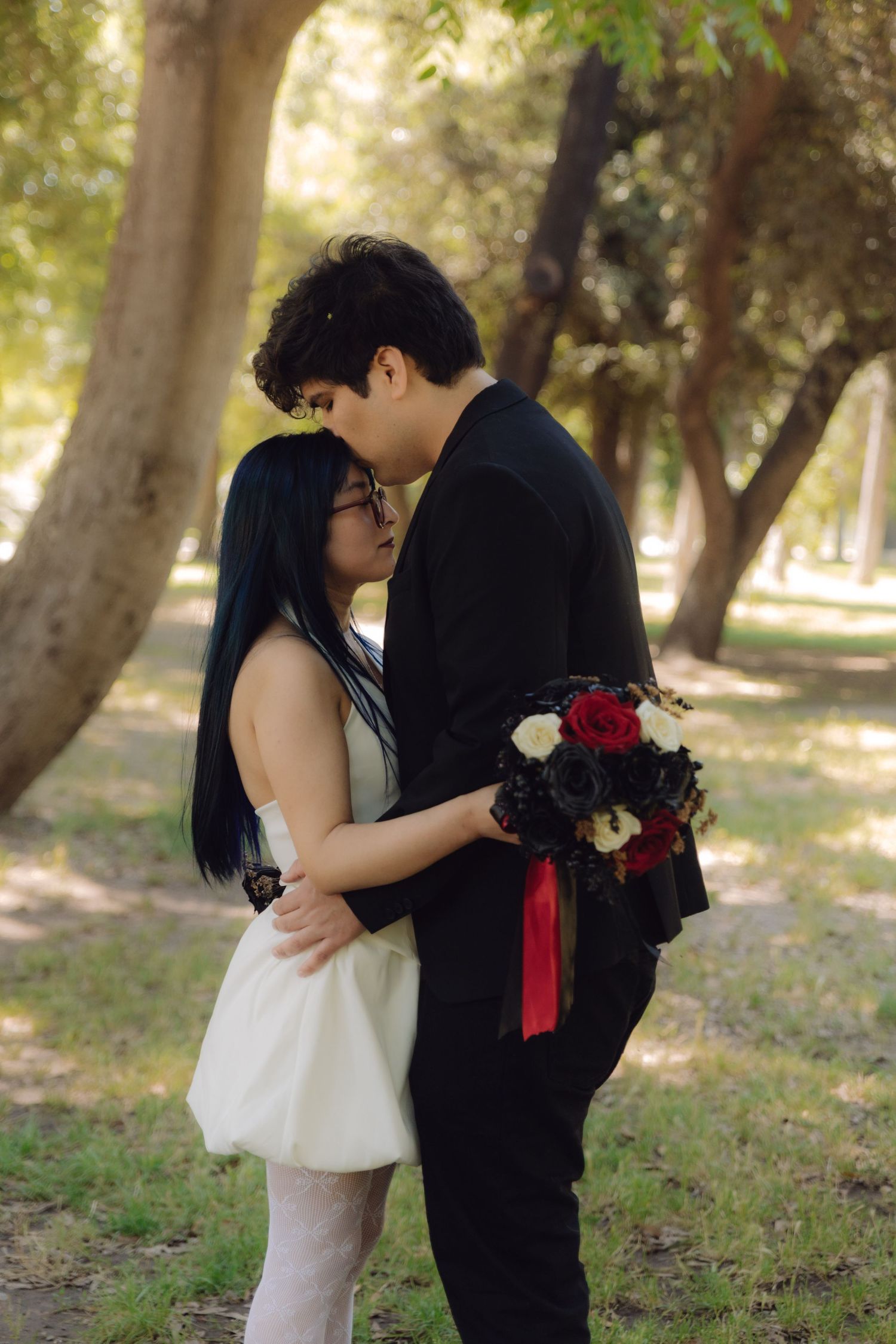Newlywed couple shares romantic moment embracing in park, groom holds red and black rose bouquet.