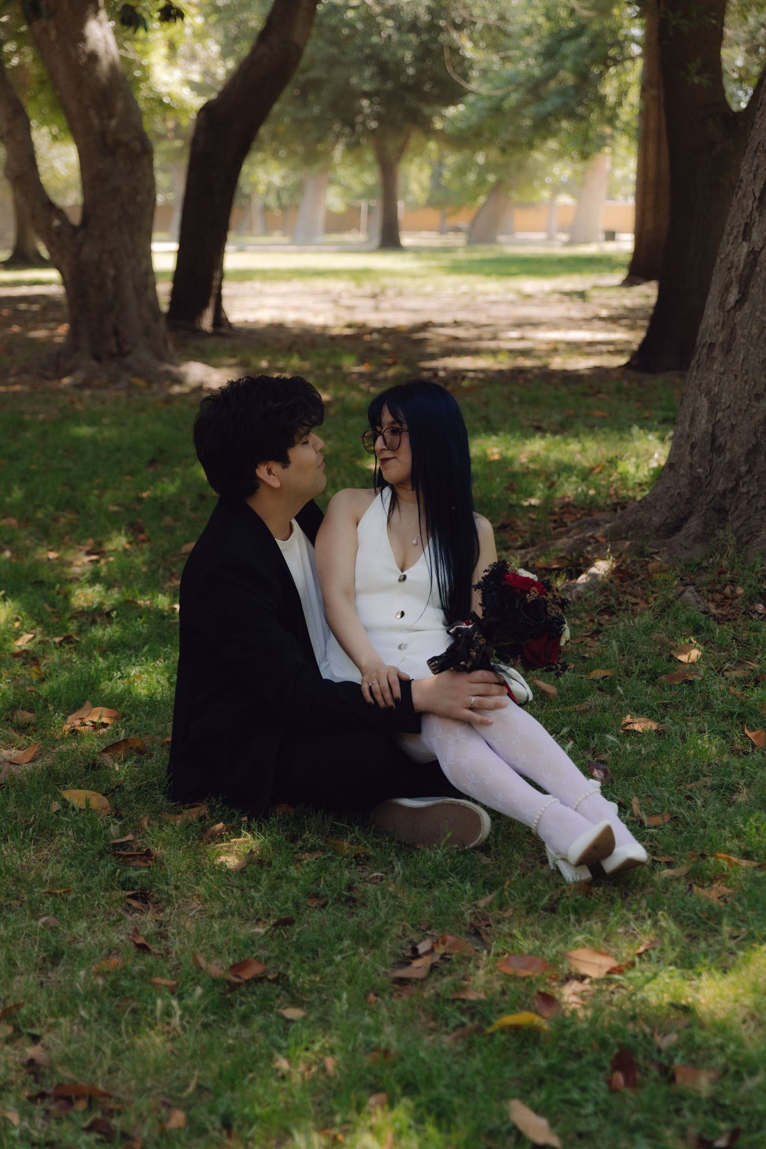 Couple sitting on grass in a park, gazing at each other, woman in white dress holding red roses.