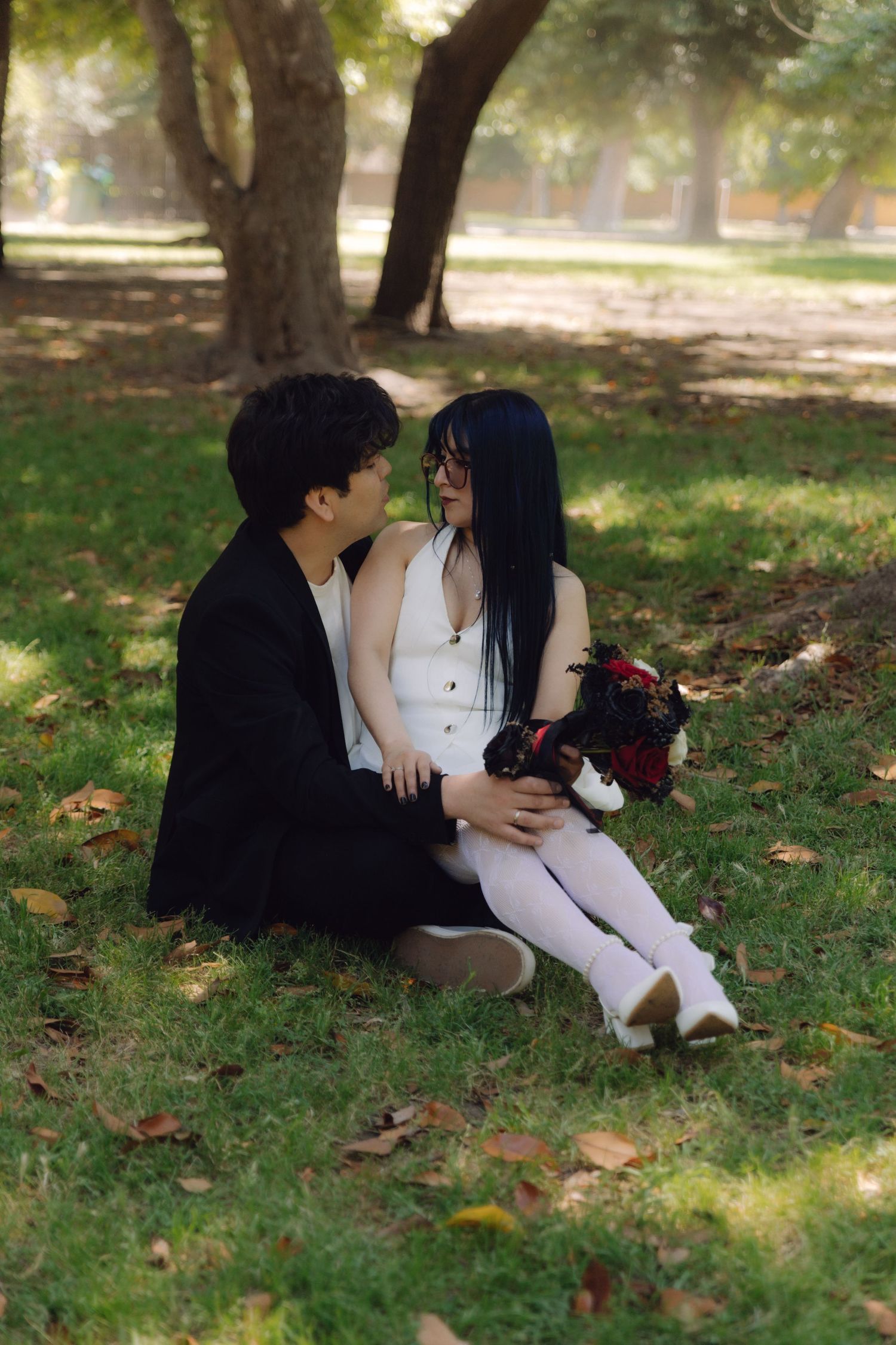 Young couple sitting together on grass under trees in a park, sharing a romantic moment outdoors.