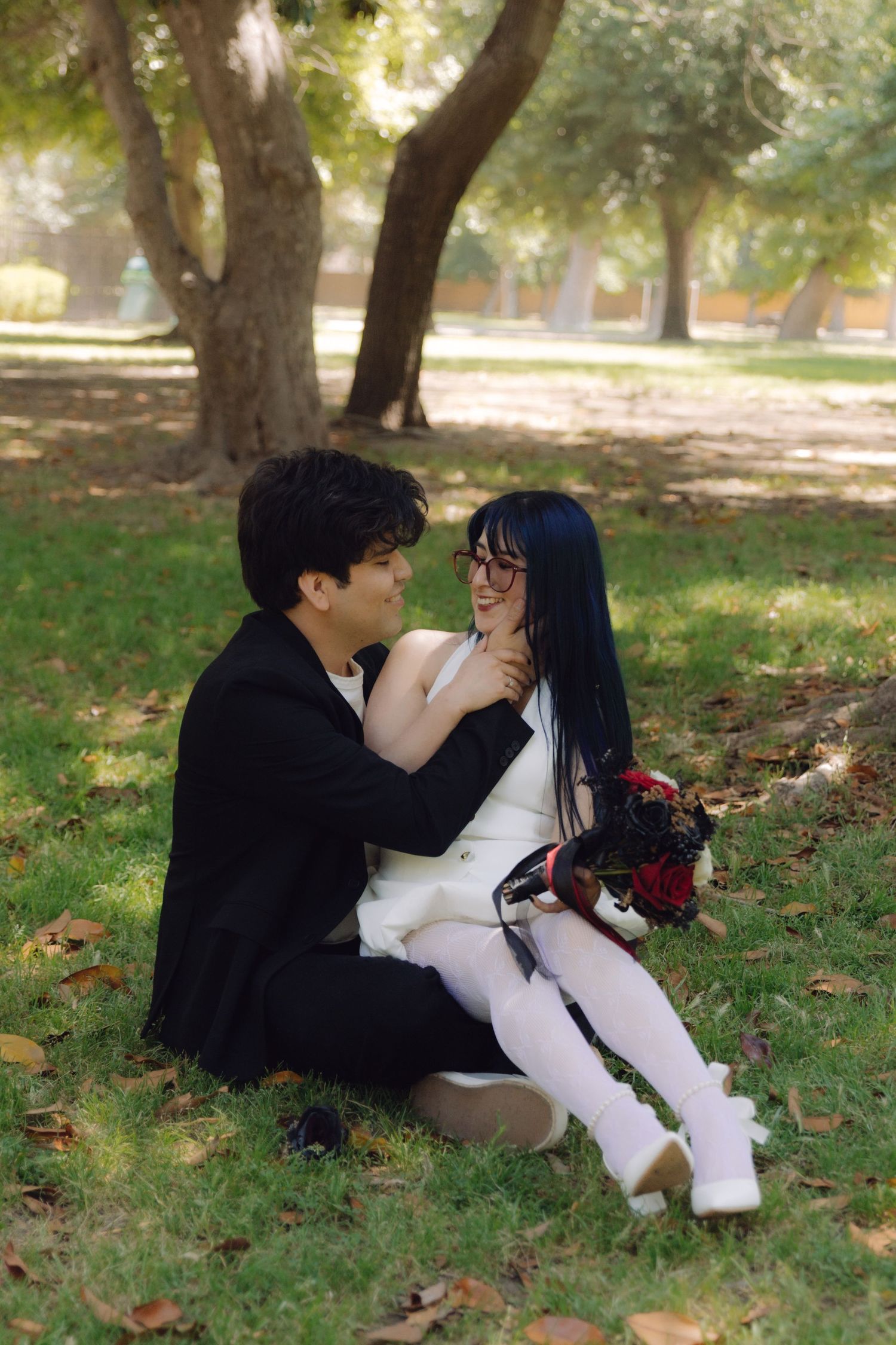 Man in black suit tenderly touches womans neck as they sit together on grass in a sunny park.