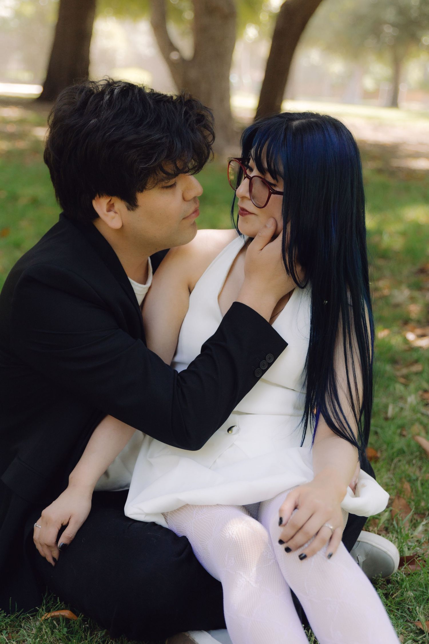Romantic couple sitting on grass in a park, man gently holding womans face as they lean close together.