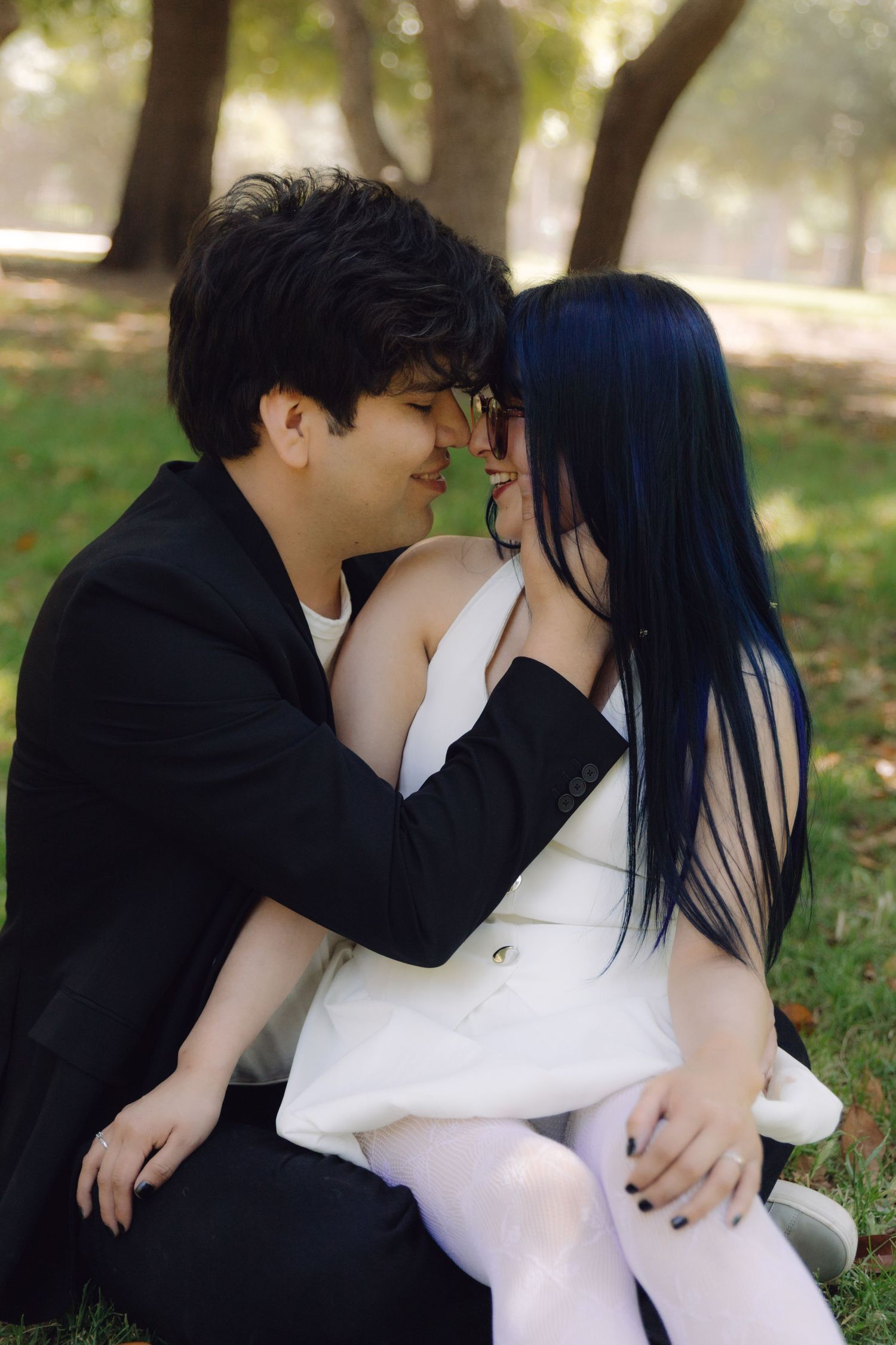 Couple sharing an intimate almost-kiss moment while sitting on grass under trees in a sunlit park.