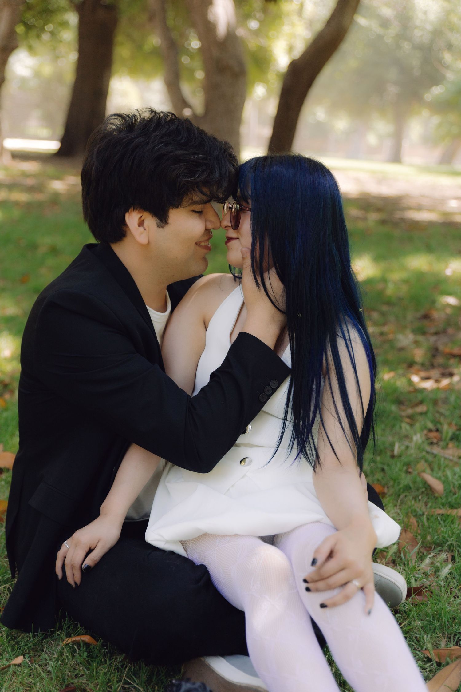Young couple in black and white formal wear share a tender moment sitting on park grass.