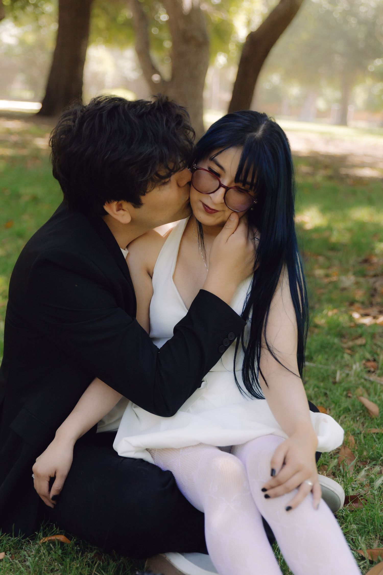 Man in black suit kissing womans cheek as she smiles while sitting together on grass in a park.