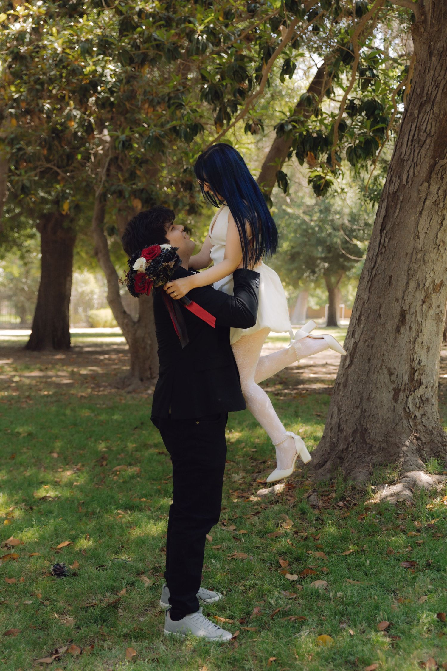 Groom lifts bride in white dress while holding red roses in a sunlit outdoor park setting.