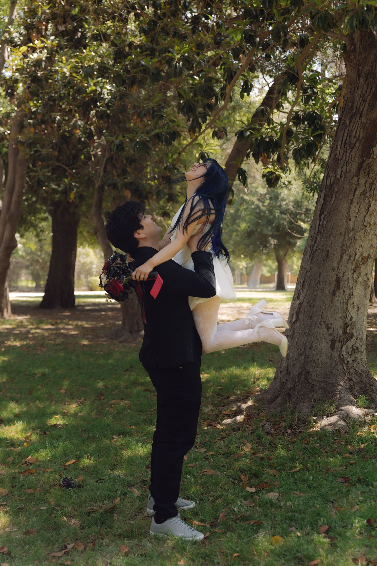 Groom joyfully lifts bride in white dress beneath a large tree in a lush green park.