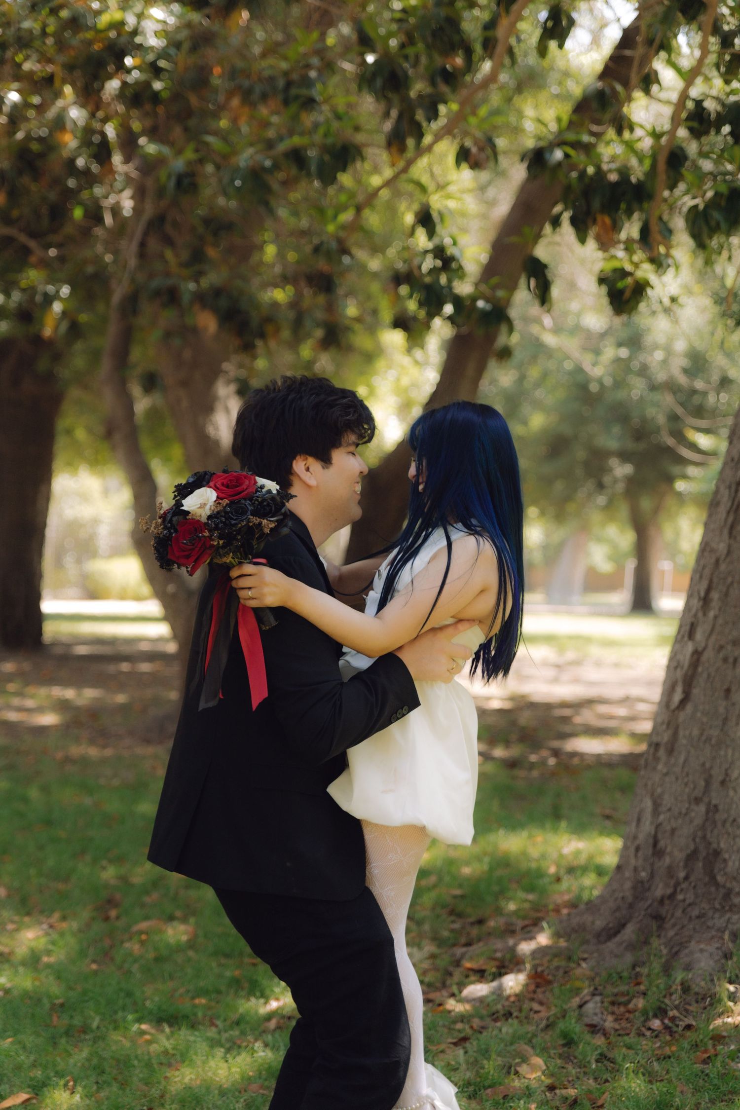 Couple in wedding attire embracing in park, bride holding red and black bouquet in white dress, groom in black suit.