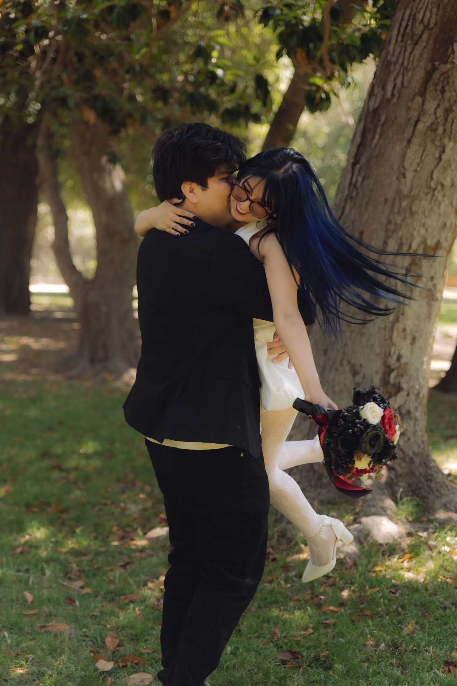Bride with blue hair kissing groom outdoors, wearing white mini dress and holding dark floral bouquet in sunny park.