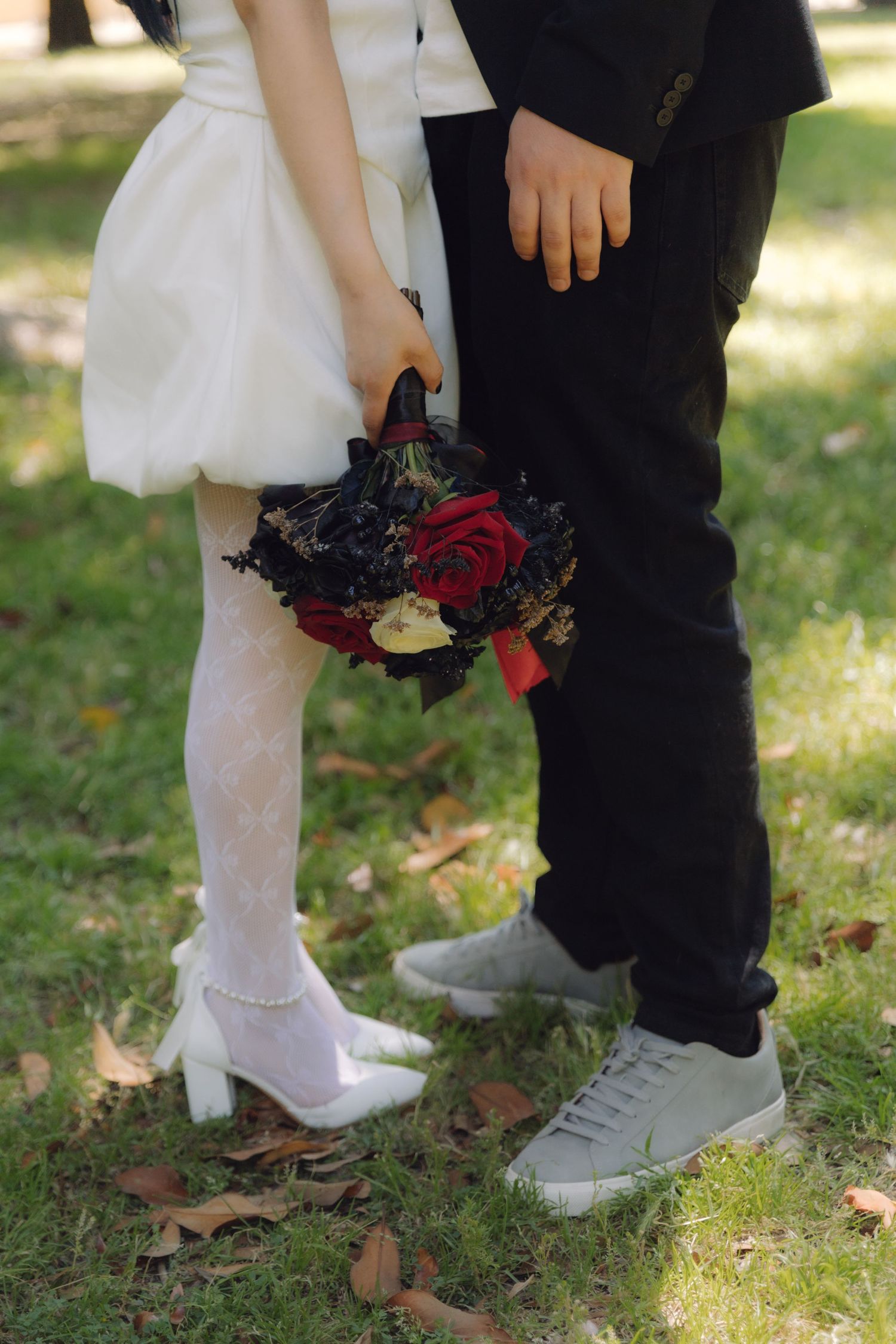 Close-up of wedding couple feet, bride in white lace tights and heels, groom in sneakers, holding gothic bridal bouquet.