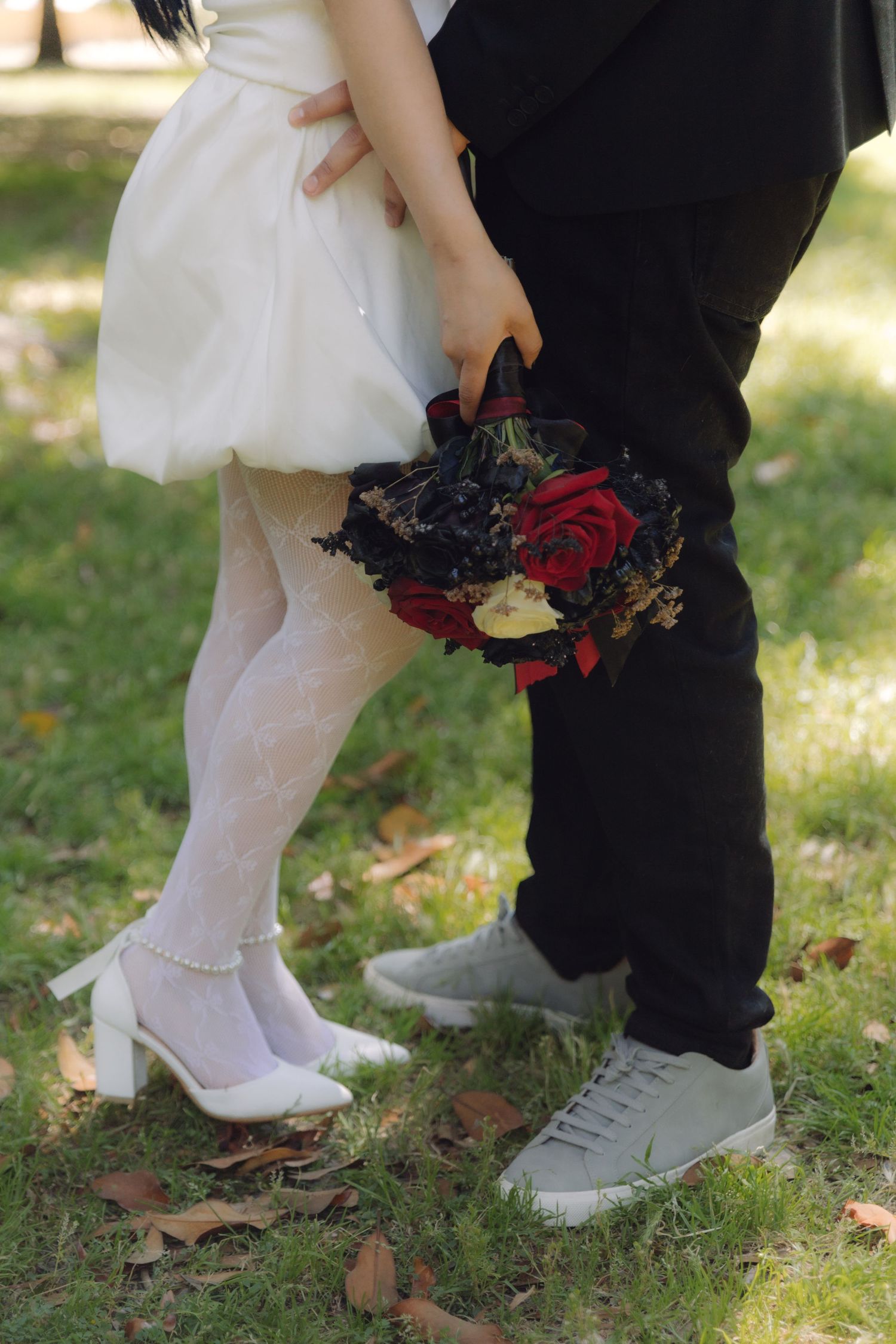 Detail shot of bride in white lace stockings and heels beside groom holding red and black rose wedding bouquet.