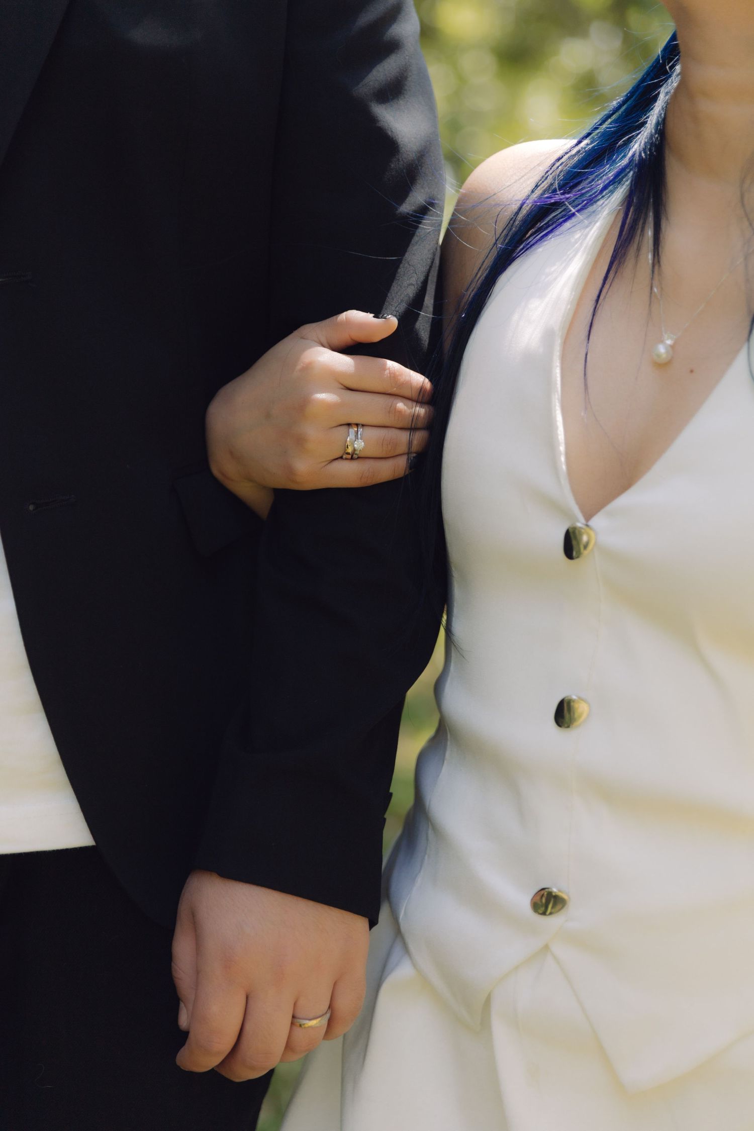 Bride in white dress with blue hair holding grooms black suit arm, both wearing wedding bands in outdoor setting.