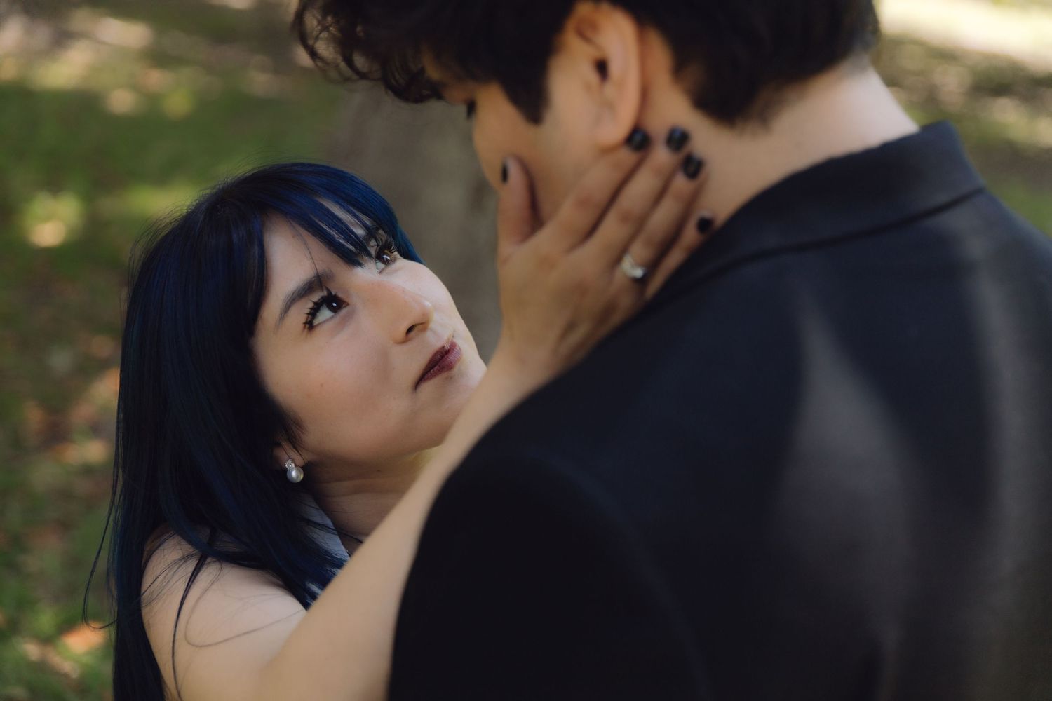 Bride with dark blue hair and black nails gazing up at groom tenderly, holding his face in natural light.