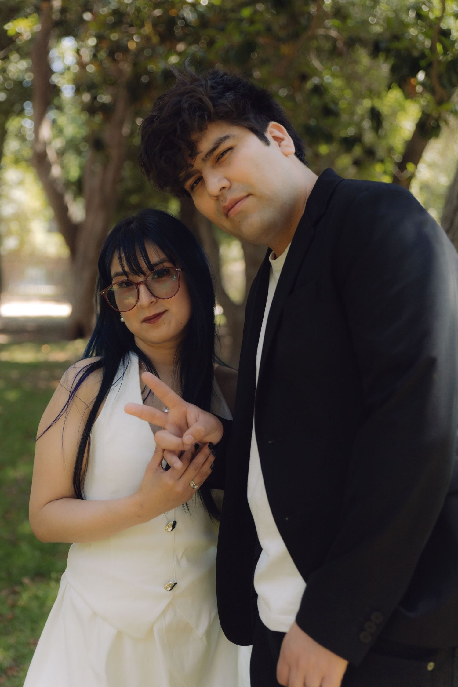 Wedding couple posing in park, bride in white dress with glasses and dark hair, groom in black suit smiling.
