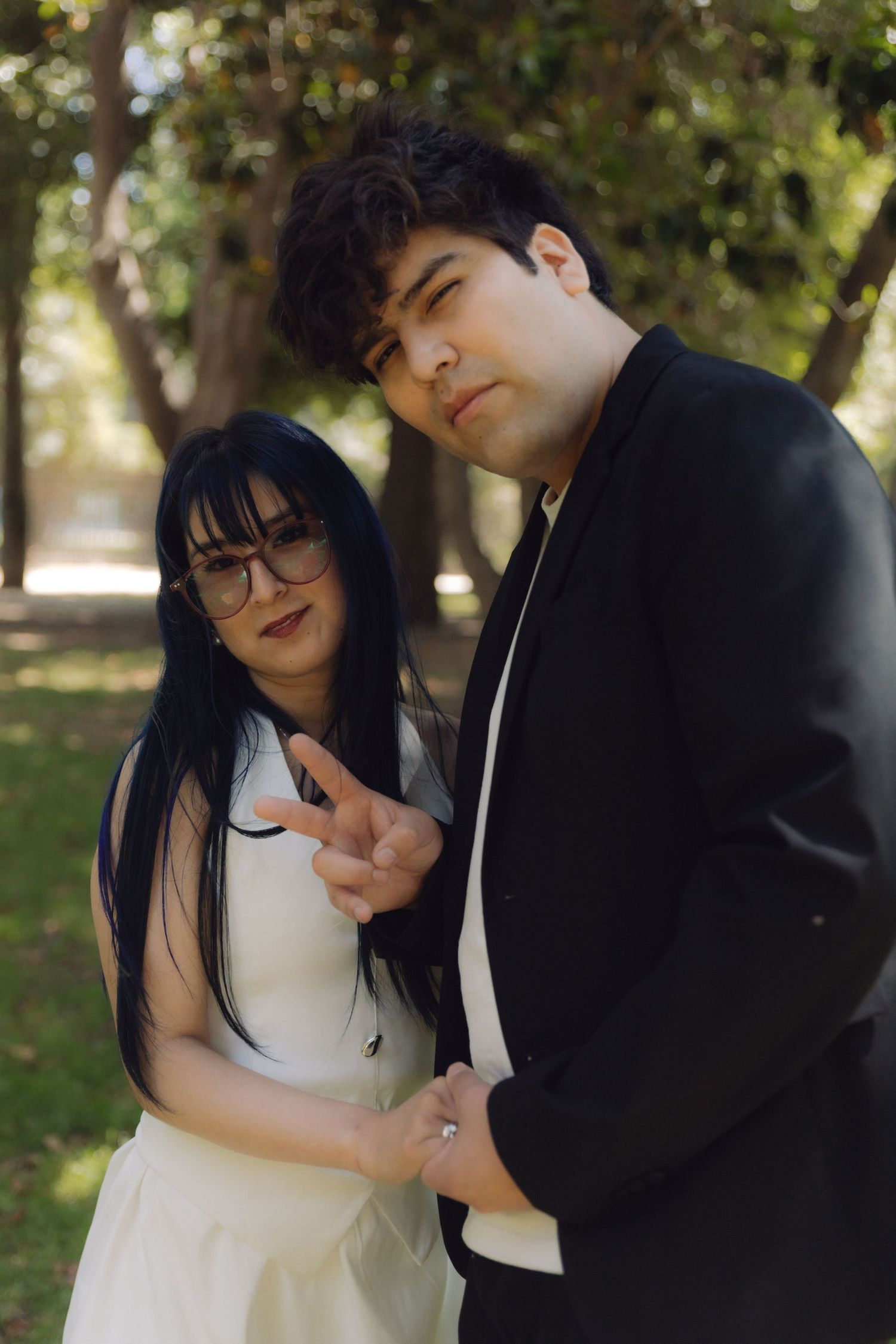Stylish wedding couple standing together outdoors, bride in white dress with glasses, groom in black suit among trees.