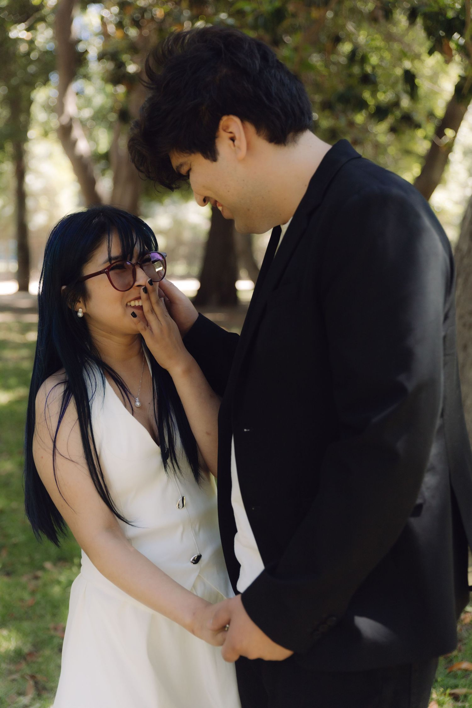 Groom tenderly touches bride's face as they stand close together in a sunlit park setting.
