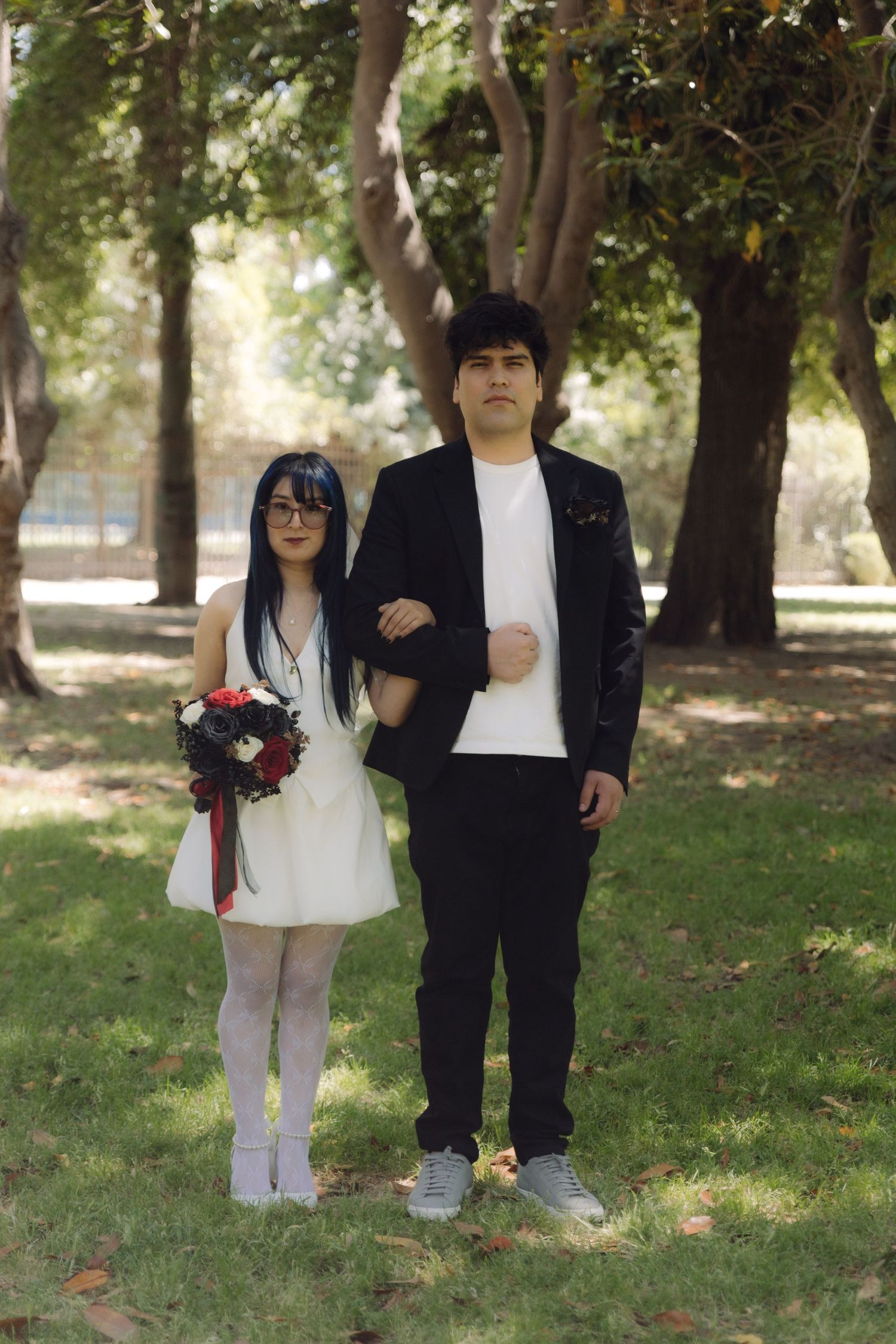 Bride and groom pose formally in park, bride holding red and white bouquet wearing white mini dress.
