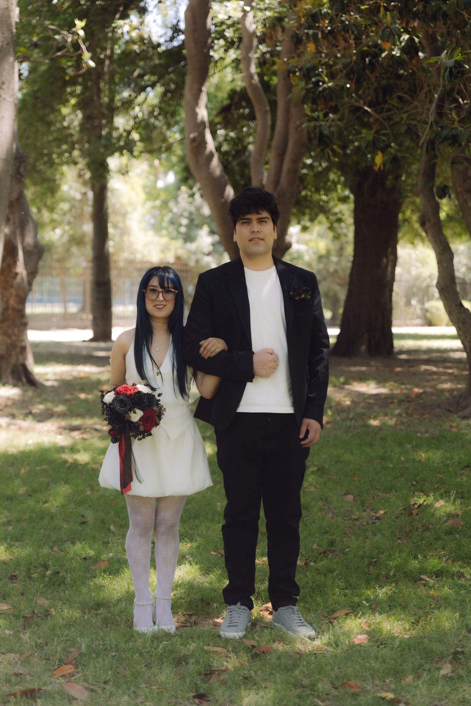 Wedding couple standing together outdoors, bride in white dress with bouquet, groom in black blazer.