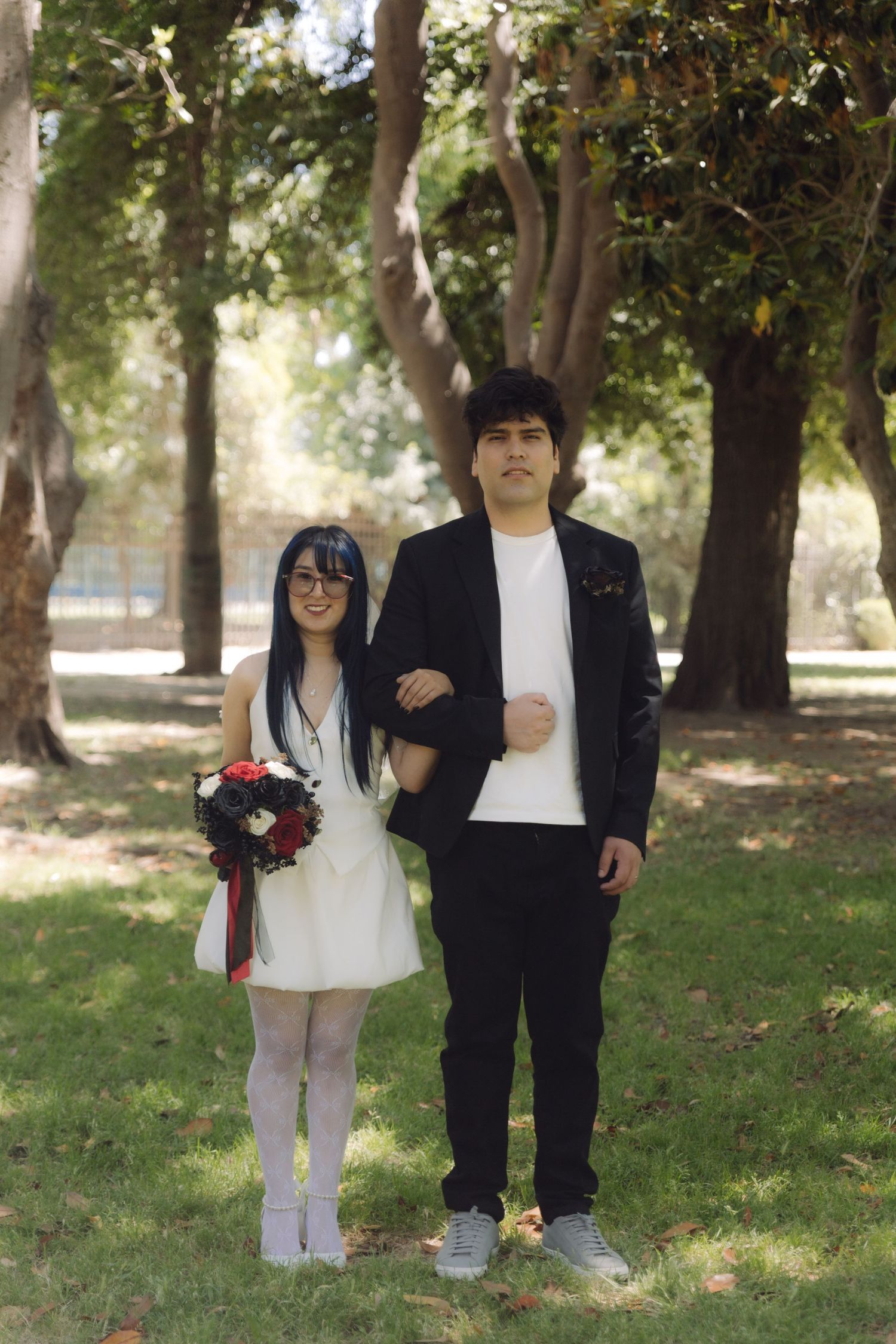 Bride and groom posing in sunny park, bride holding floral bouquet in white dress and white tights.