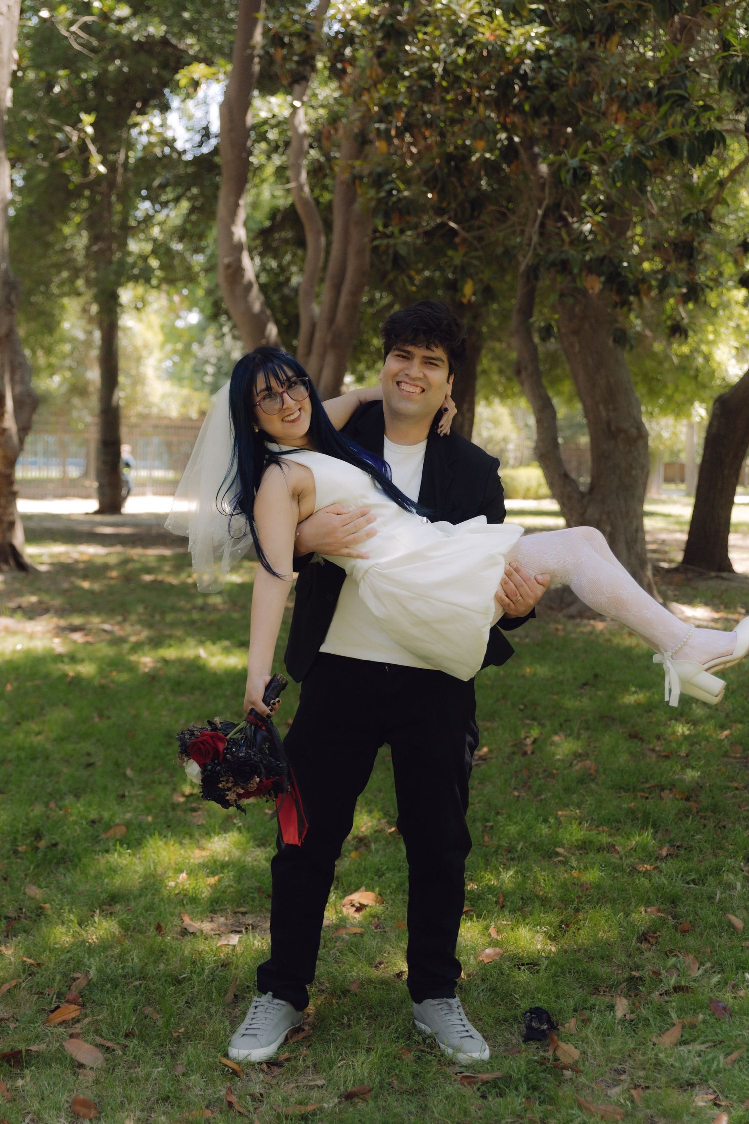 Groom carries smiling bride in his arms outdoors in a park on their wedding day.