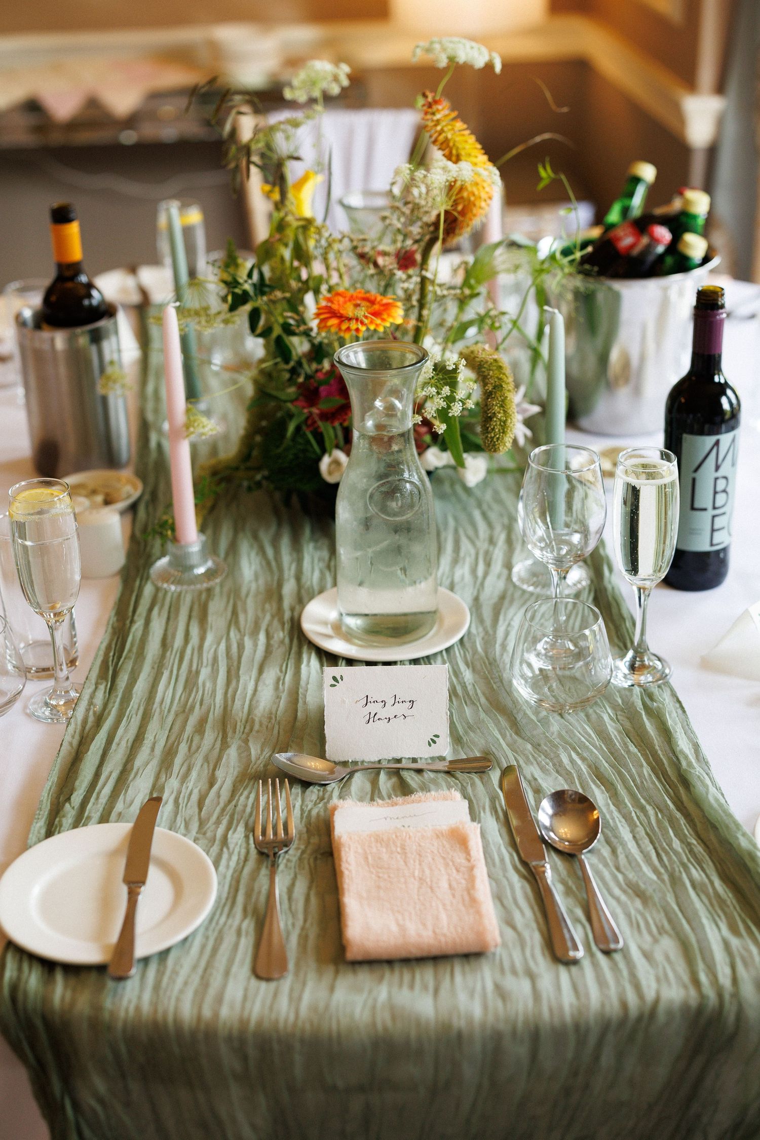 Smiling newlyweds showing wedding rings at outdoor reception table with floral centerpiece and green trees.