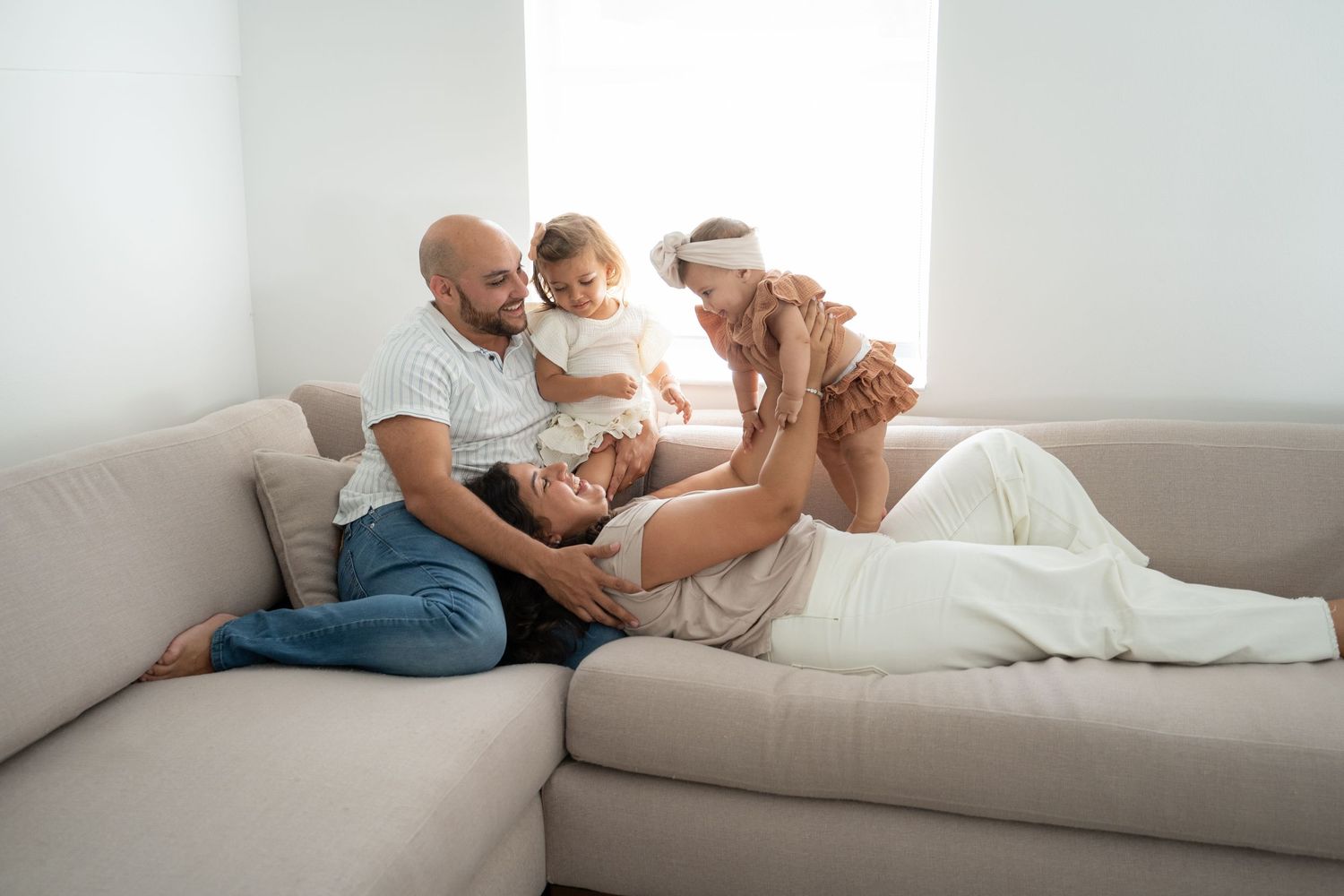 Happy family relaxing on a cozy beige sofa, parents playing with their toddler in a bright modern living room.