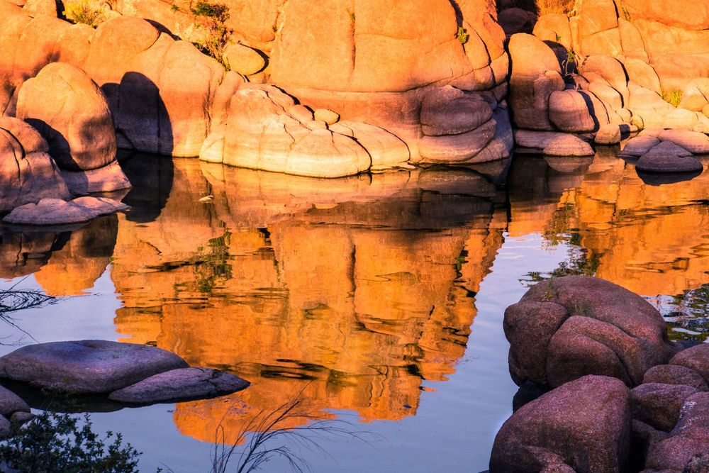 Golden orange rocks reflect in still water at sunset, creating a mirror-like scene in a desert canyon landscape.