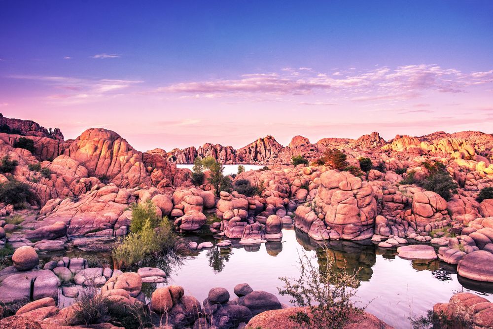 Pink granite boulders surround a tranquil desert pond at sunset in a rocky landscape with lush green shrubs.