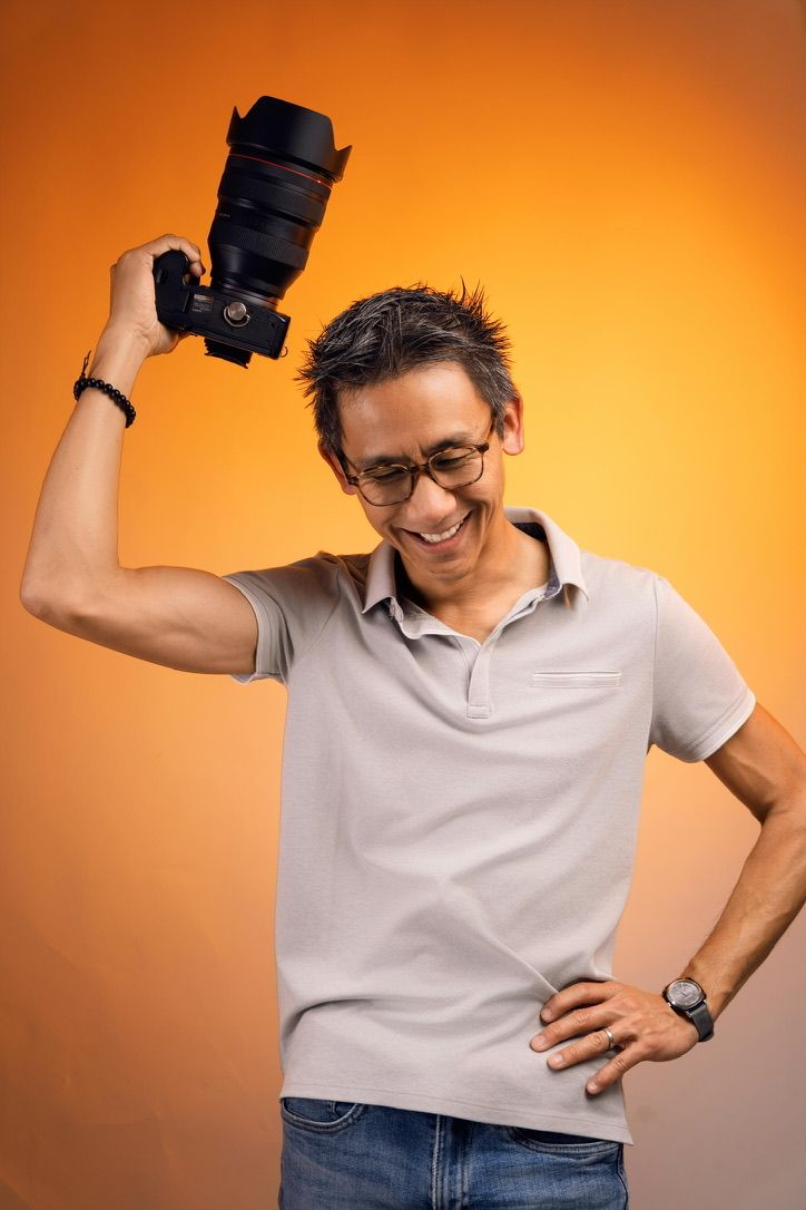 A man in glasses holds a DSLR camera up to his eye against an orange background, wearing a gray polo shirt.
