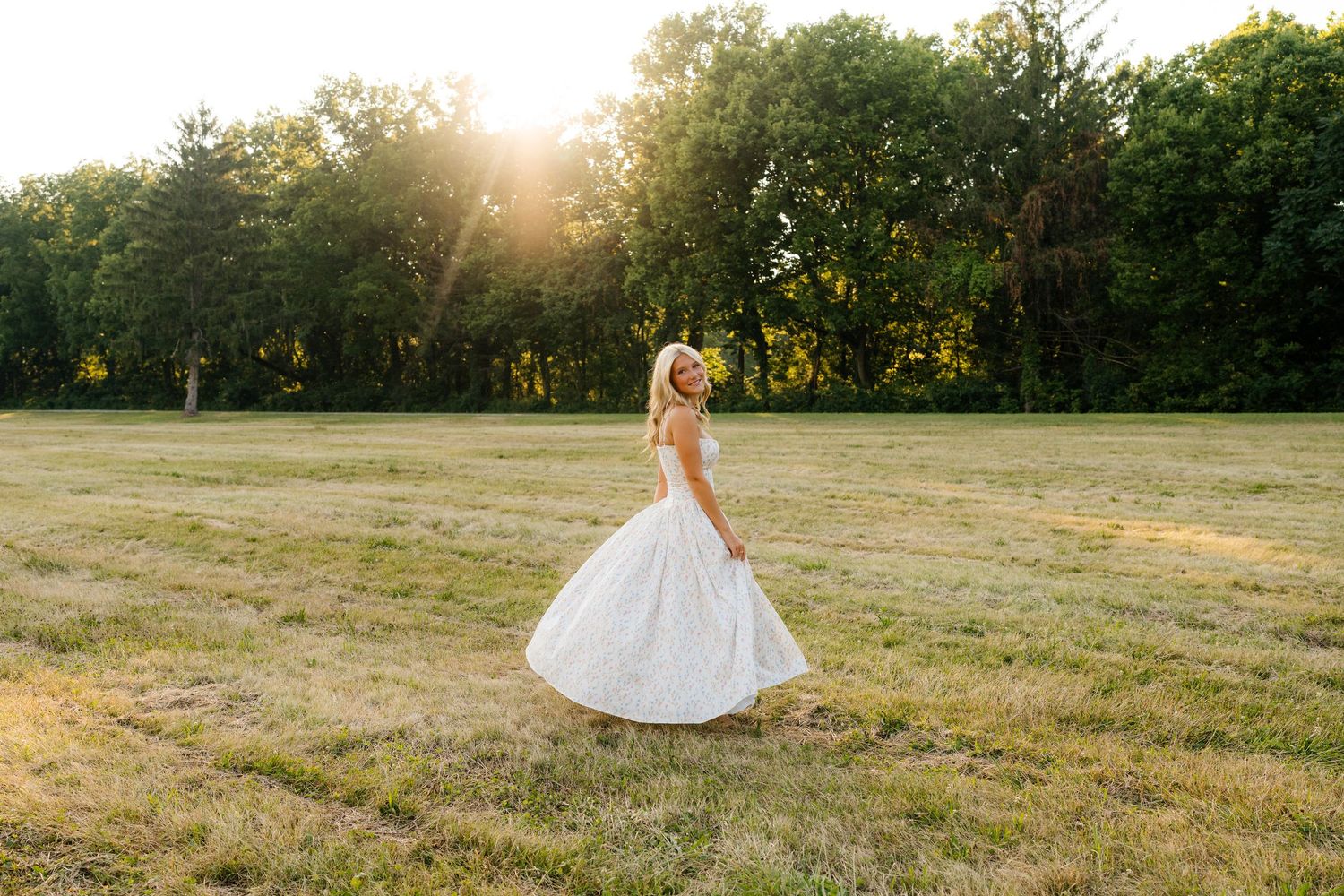 A blonde woman in a flowing white lace dress walks through a sunlit green field at golden hour.