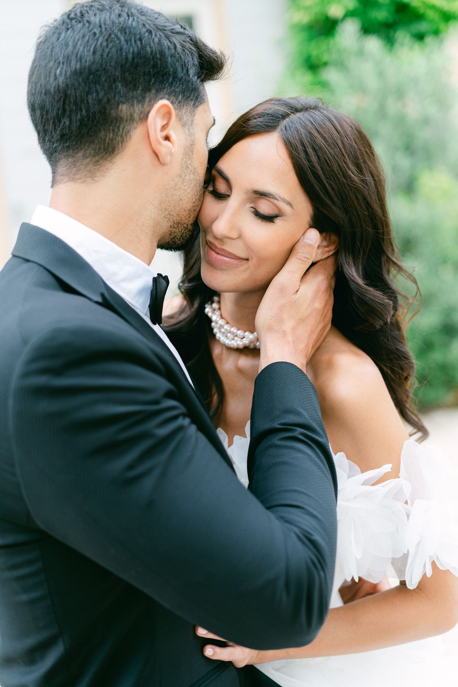A bride and groom share a romantic kiss outdoors, with the groom in a black tuxedo and bride in a white ruffled gown.