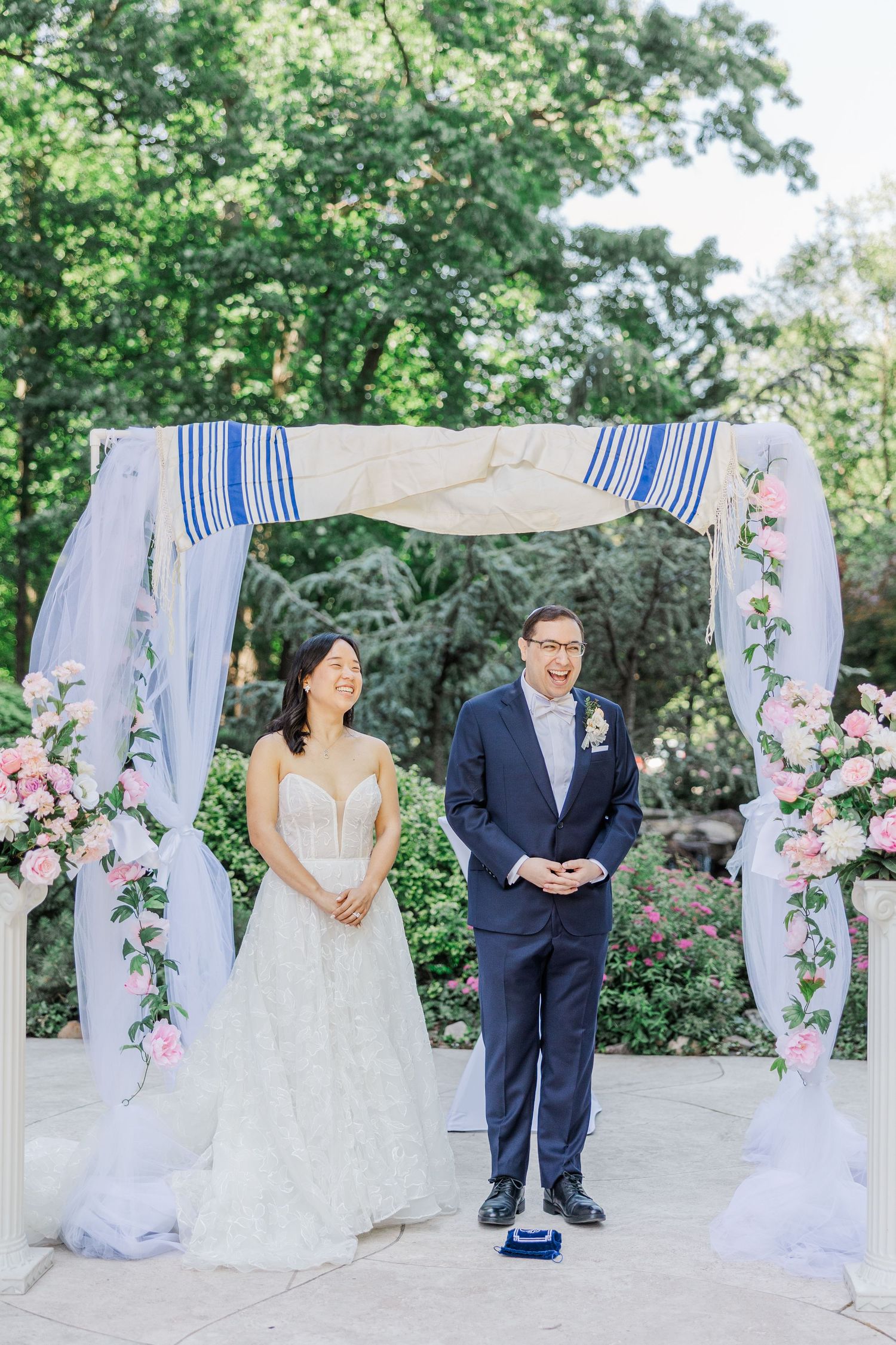 Bride and groom laughing under a floral chuppah with pink roses and blue striped tallit at an outdoor wedding ceremony.
