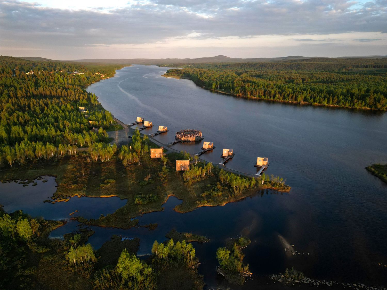 Aerial view of floating cabins on a serene river surrounded by lush green forests at golden hour.