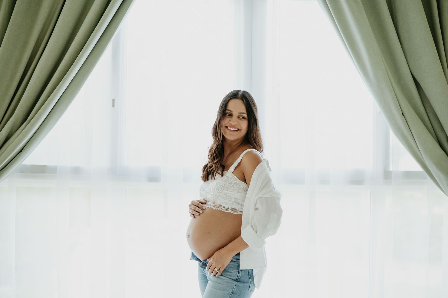 Smiling pregnant woman in white lace bralette and jeans cradling baby bump near elegant sage green curtains.