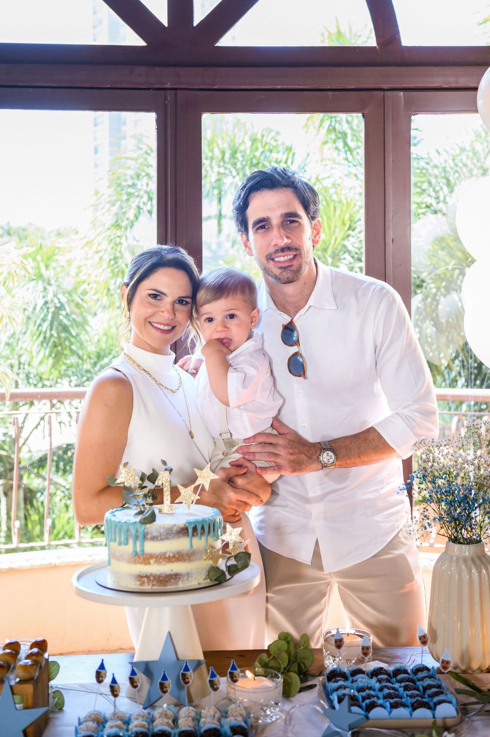 Family celebrating baby's first birthday with a decorated cake and dessert table at an outdoor party venue.