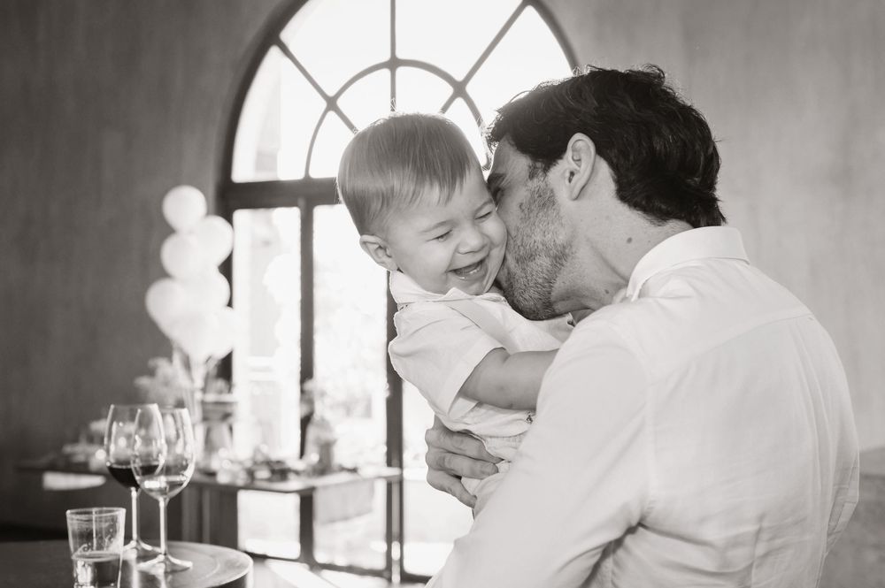 Black and white photo of a father kissing his laughing toddler son at a family celebration indoors.